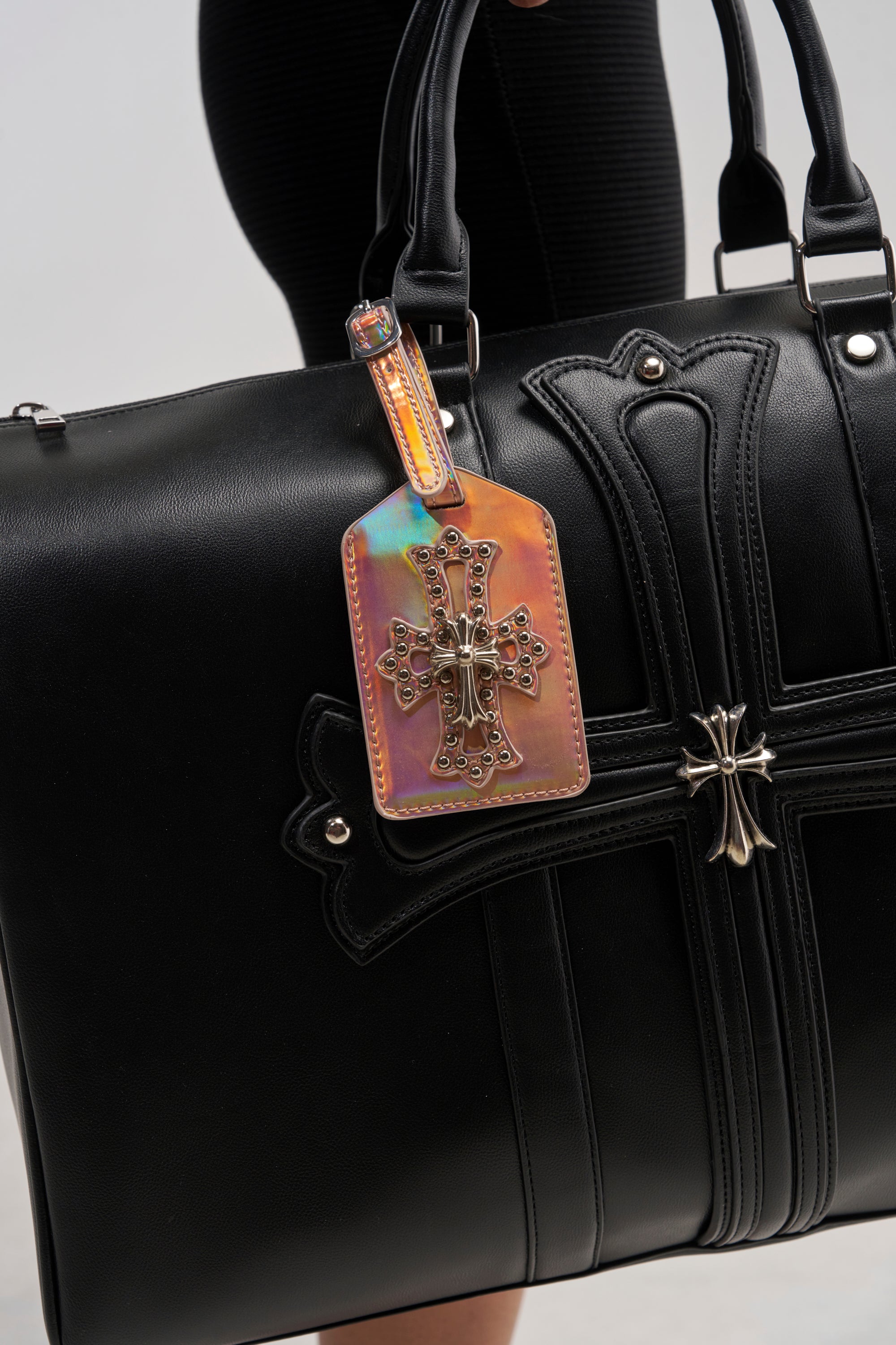 A person holds a black leather bag with cross designs, featuring the FEELIN ROSIE LUGGAGE TAG—a metallic, multicolored tag adorned with a cross and metal studs—hanging elegantly from one handle.