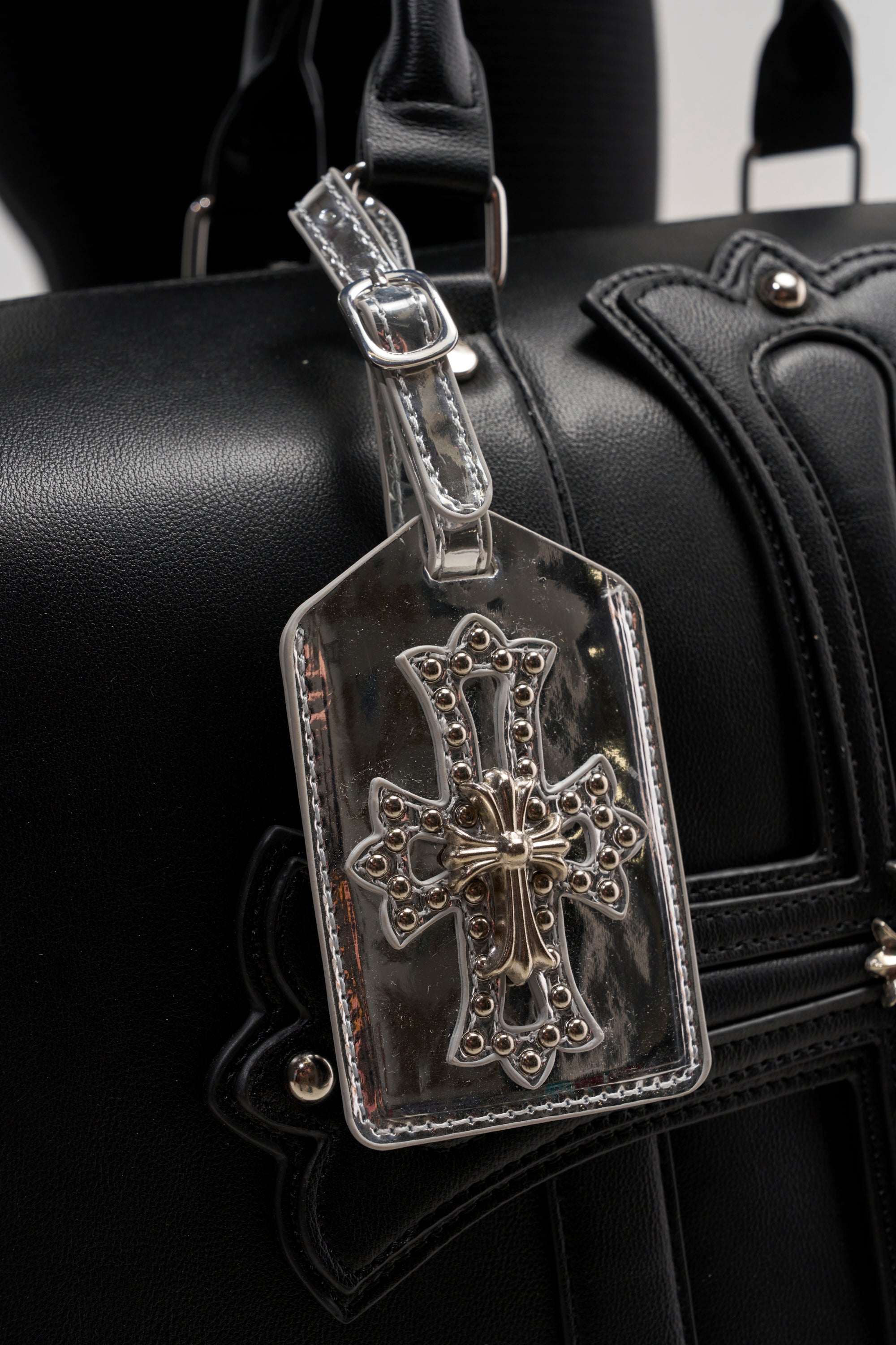 The black leather bag is accented by the IM CHROME LUGGAGE TAG, which is crafted from shiny silver and showcases an ornate cross design. The detailed embellishments and raised patterns on the tag give it a striking and decorative look.