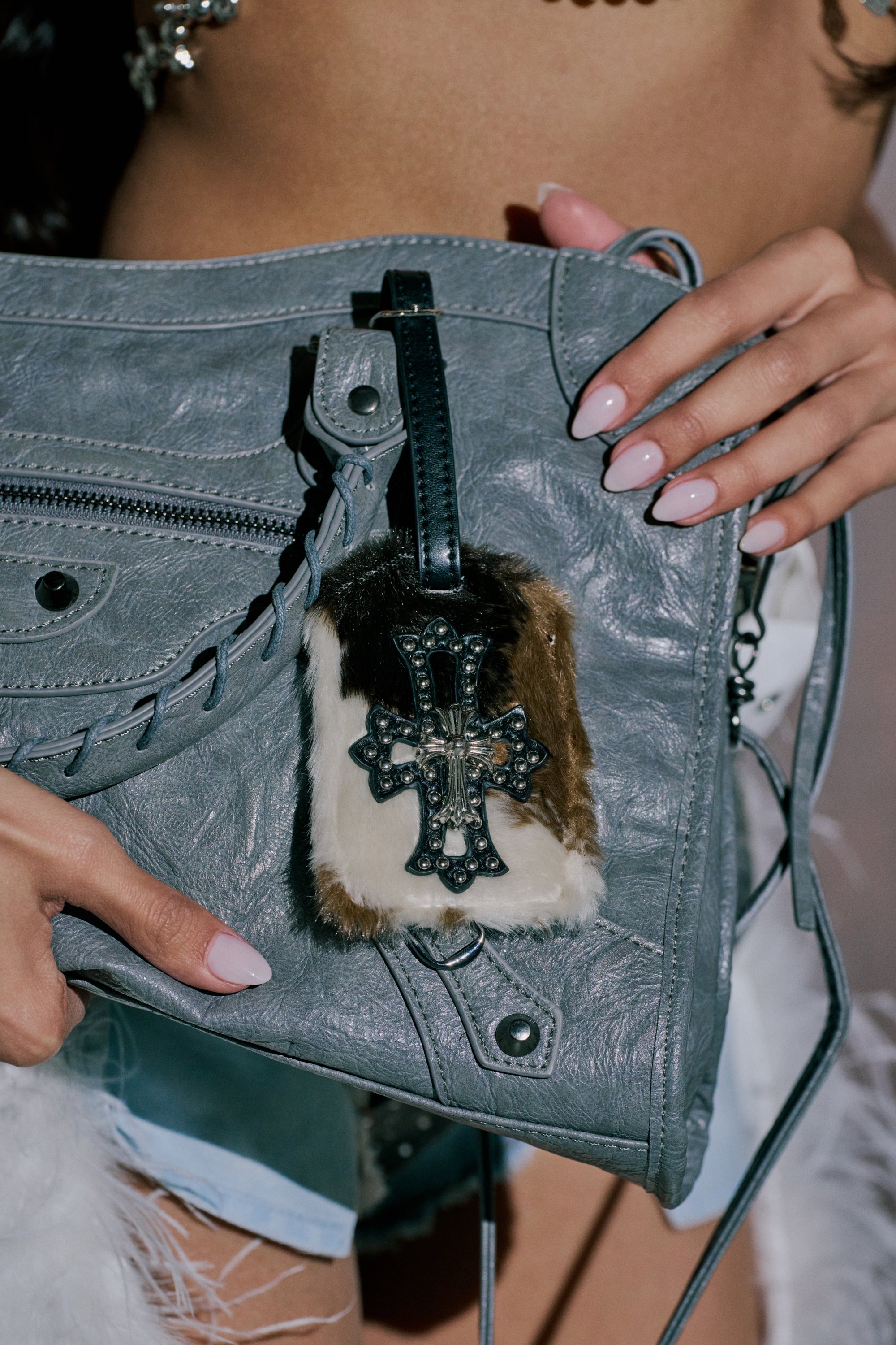 A person with manicured nails holds a gray leather bag featuring the HIGH STANDARDS FAUX FUR LUGGAGE TAG, which is adorned with an ornate metal cross. Their bare torso and some blue clothing appear in the background. 0