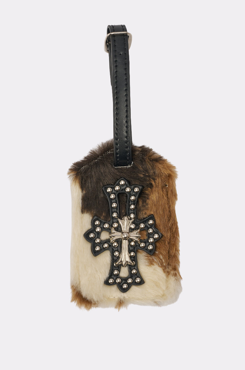 The HIGH STANDARDS FAUX FUR LUGGAGE TAG features brown, white, and tan cow-print faux fur, a black leather strap, and a studded metal cross—making it a standout travel accessory. 1