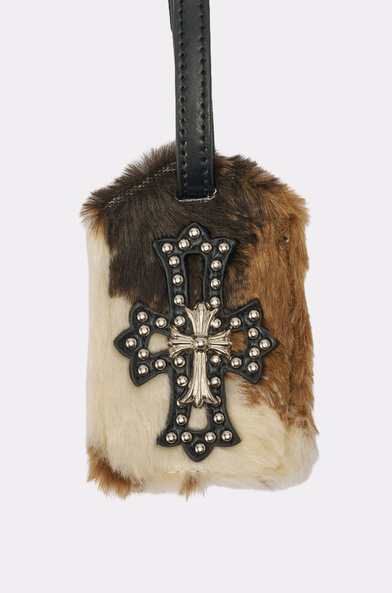 The HIGH STANDARDS FAUX FUR LUGGAGE TAG is a stylish brown and white cow-print accessory with a black faux leather strap, featuring a decorative metal cross and bead accents on the front. 3