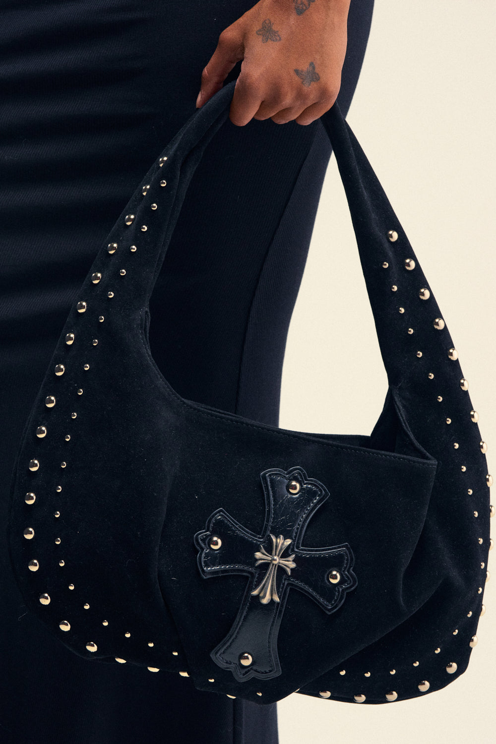 A person holding the REALLY THAT GIRL SUEDE BAG, an oversized shoulder bag made from black faux suede and embellished with silver studs and a cross patch with metal detailing. Their tattooed hand rests on the bag as they wear a dark, ribbed outfit.