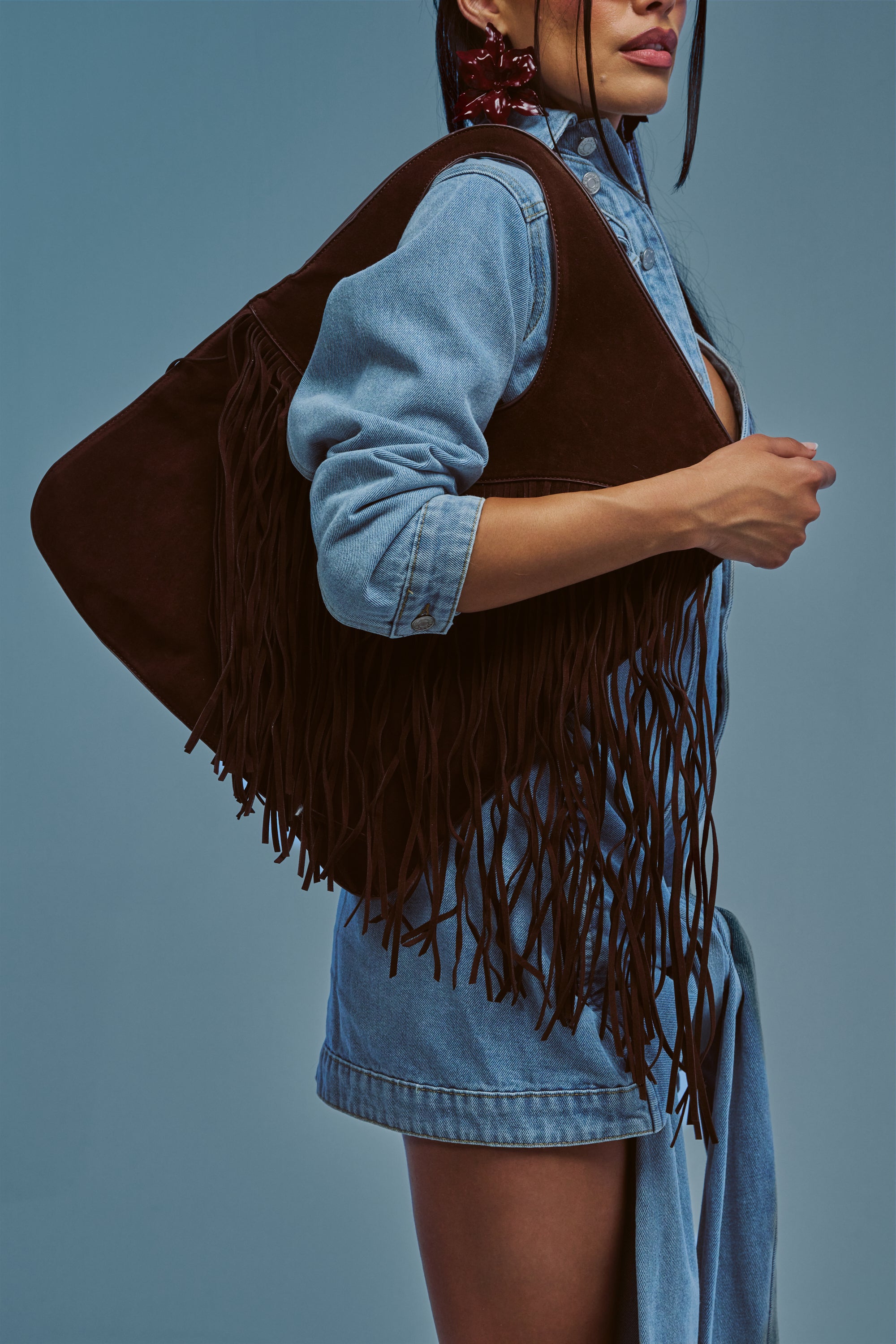 A woman in a denim dress and brown fringed vest poses against a solid blue background, standing sideways with one hand on her waist and her long dark hair partly obscuring her face. She finishes off her boho-inspired ensemble with the FRINGED OUT SHOULDER PURSE, giving the look an added dose of casual chic.