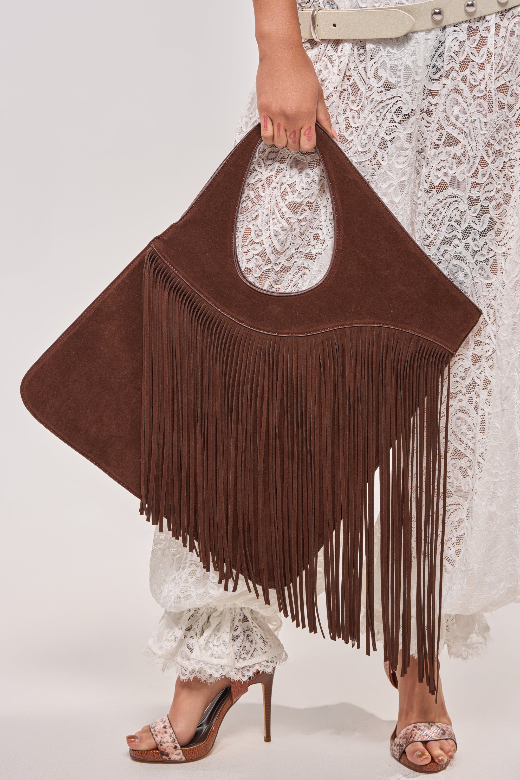 A person in a white lace outfit and high-heeled sandals holds the FRINGED OUT SHOULDER PURSE, a large boho-chic bag with long western fringe details and a rounded handle. 1
