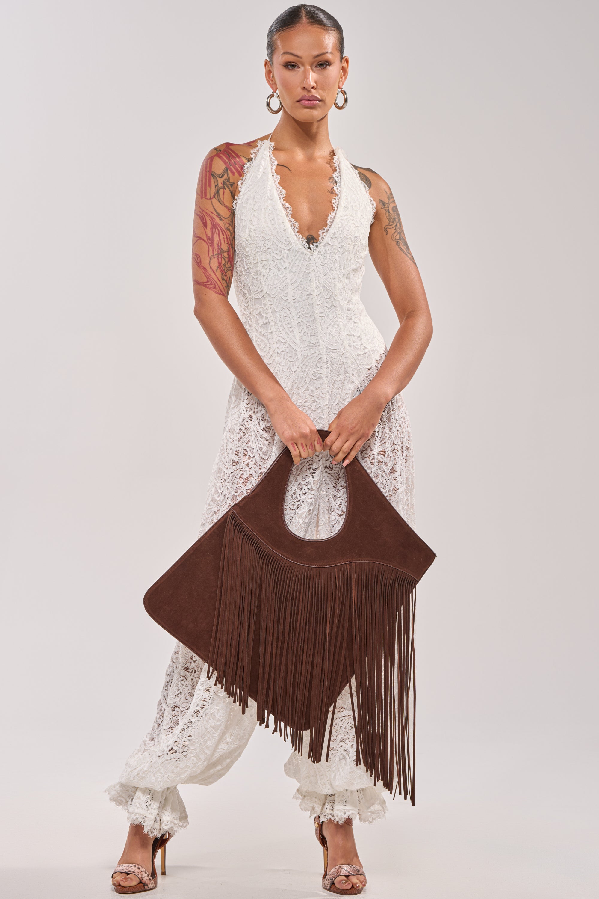 A woman stands holding the FRINGED OUT SHOULDER PURSE, dressed in a sleeveless white lace jumpsuit with frilled ankles, hoop earrings, high heels, and a right arm tattoo. Her hair is slicked back. 2