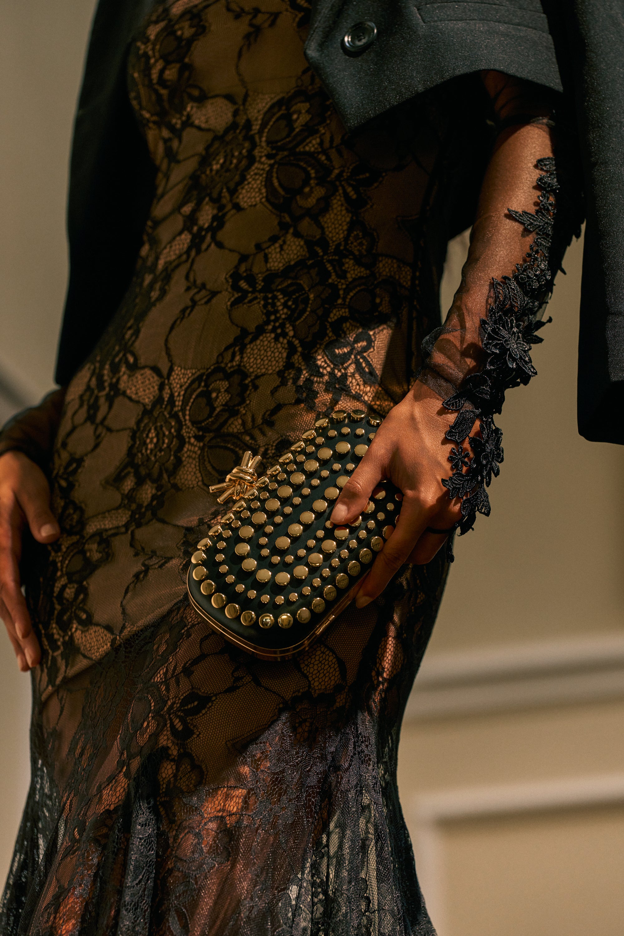 A person in a sheer black lace dress holds the STUDS FOR LIFE PURSE. The dress features floral lace patterns and intricate sleeve details, while a dark jacket is draped over one shoulder.