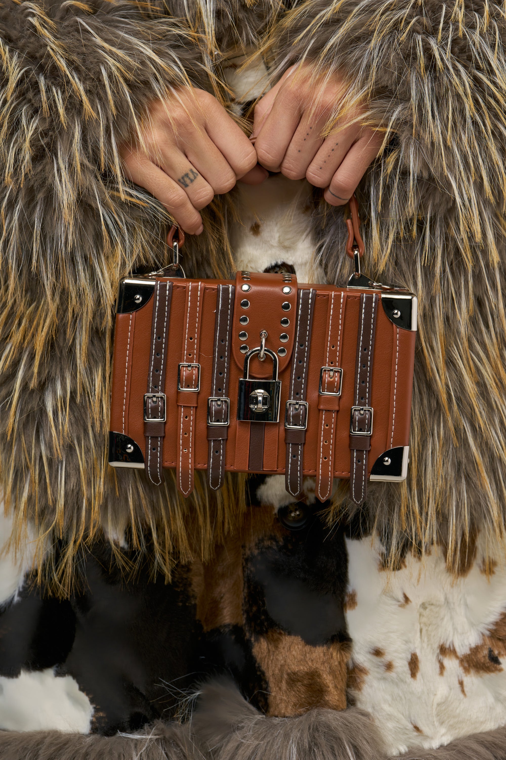 A person holds a LOCK AND STRAP BAG, a brown, boxy trunk-style streetwear handbag with silver metal corners, multiple buckle-embellished leather straps, and a central padlock clasp.