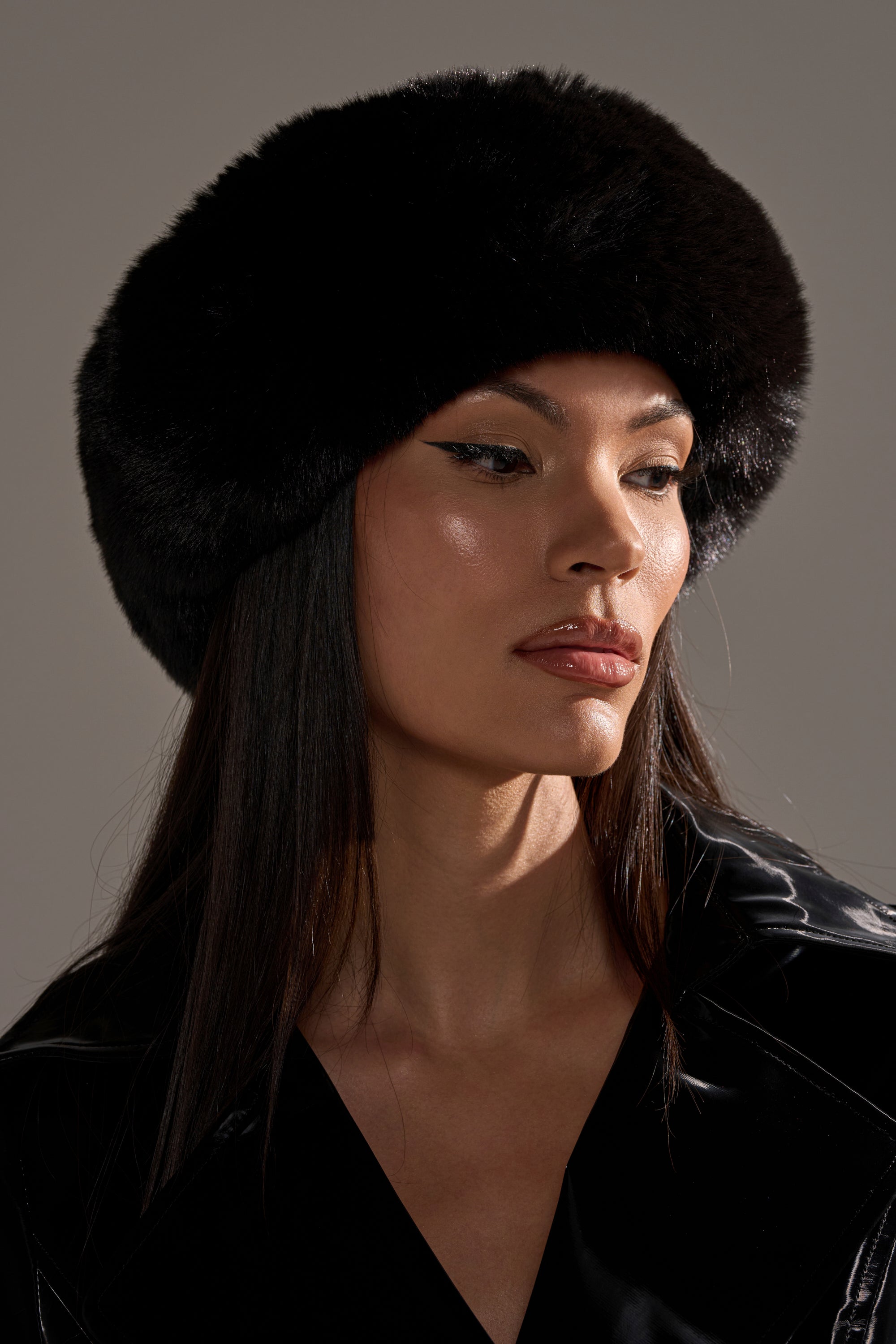 A woman with straight dark hair wears the LUXE JENNY FAUX FUR HEADBAND IN BLACK and a matching outfit. With winged eyeliner and glossy makeup, she gazes to the side, showcasing a chic winter accessory against a neutral background.