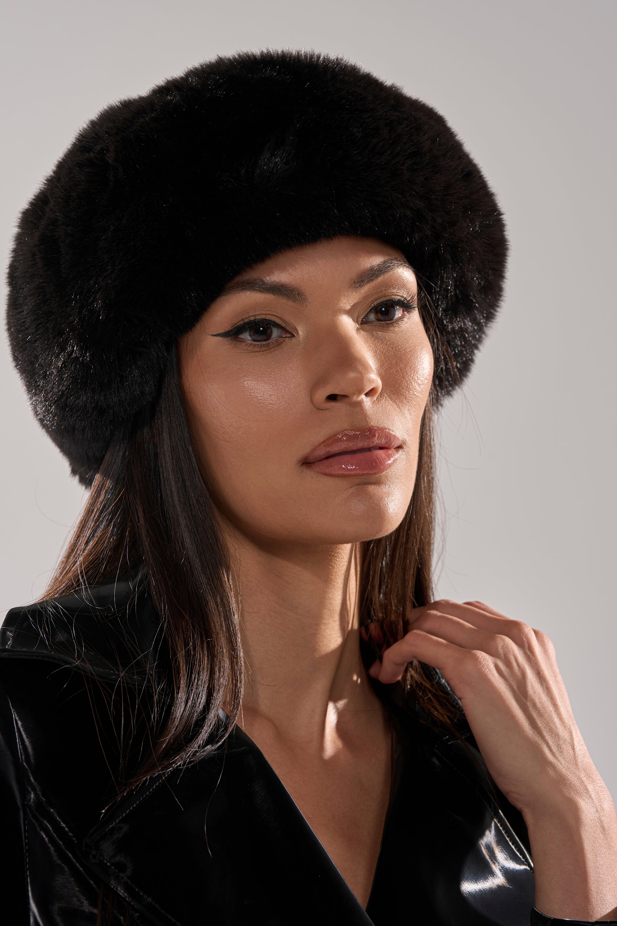A woman with long dark hair wears the LUXE JENNY FAUX FUR HEADBAND IN BLACK and a shiny black coat, gazing ahead with a neutral expression—an elegant display of winter style against a minimalist background.