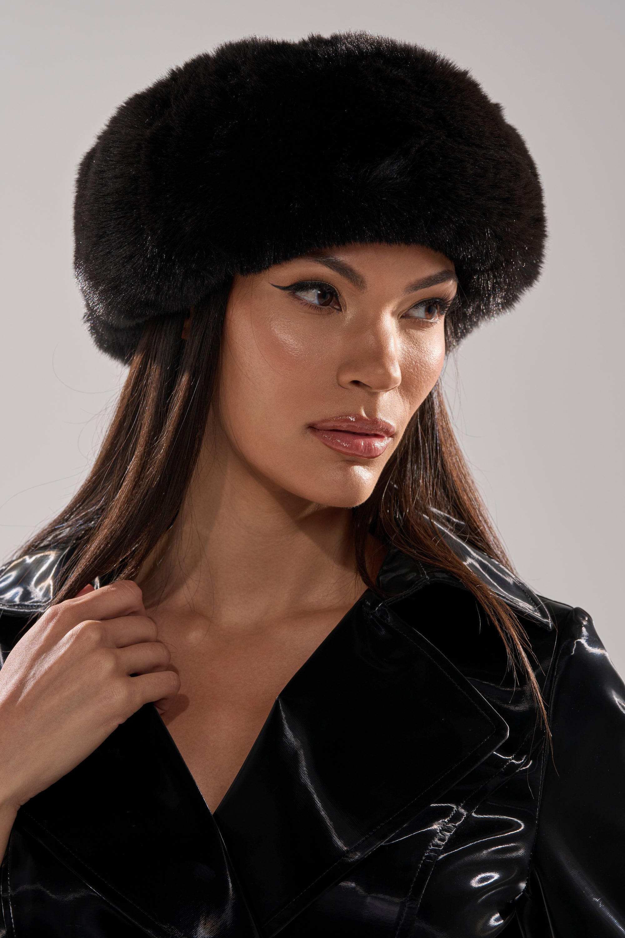 A woman models the LUXE JENNY FAUX FUR HEADBAND IN BLACK with a shiny black jacket, her long brown hair visible beneath the accessory. The light background highlights her chic winter style.