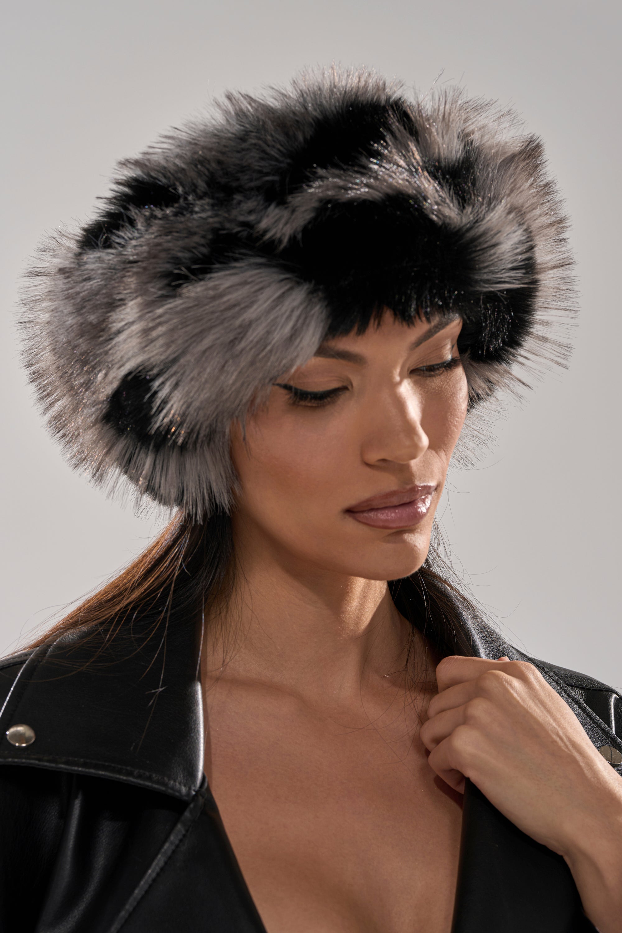A woman in a black leather jacket wears the ARLO FAUX FUR HEADBAND, featuring fluffy black-and-gray faux fur and satin lining, looking downward with a soft expression against a neutral background. 0