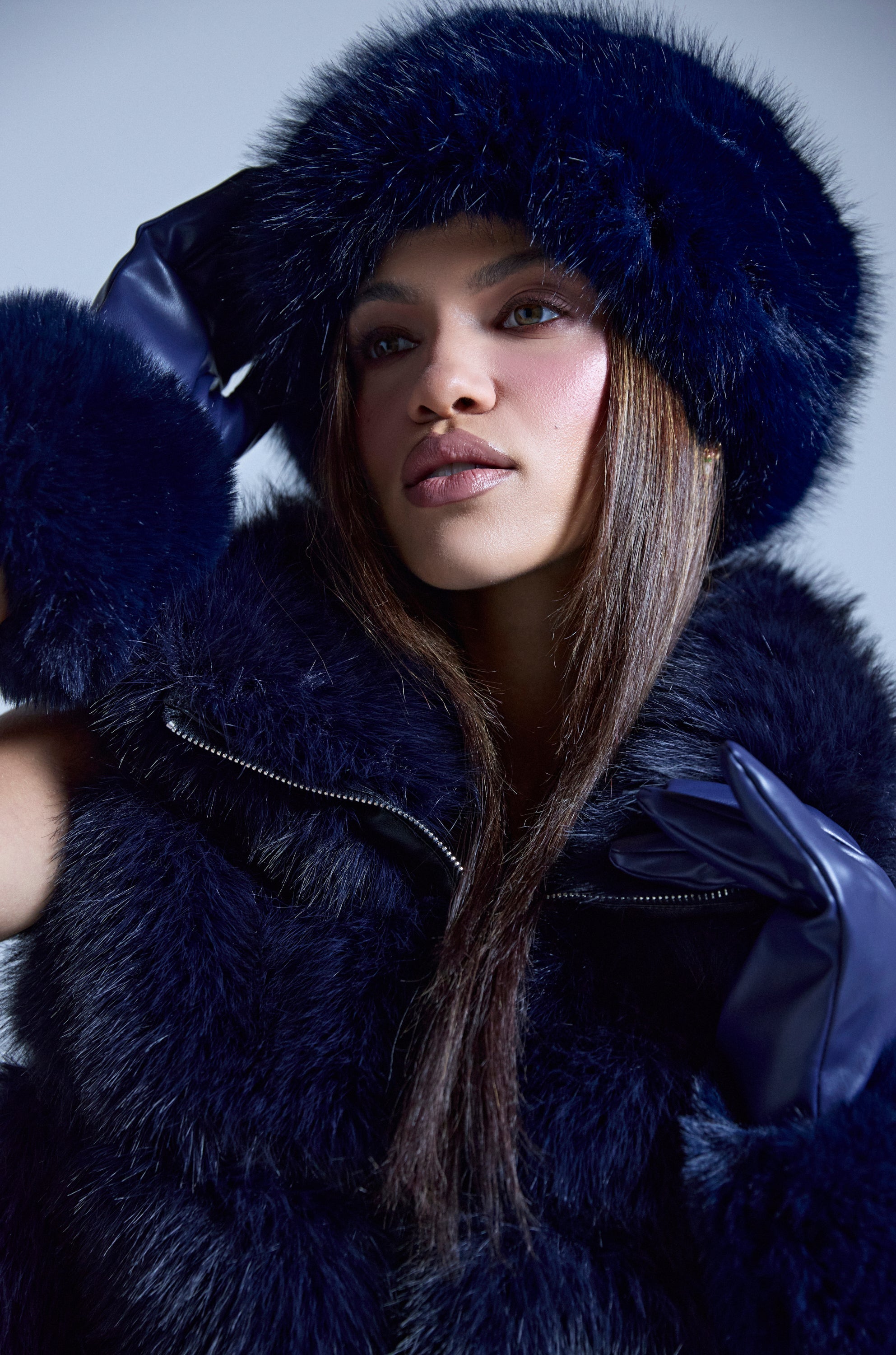A woman with long straight hair and a neutral expression poses against a light background, wearing the SO CHIC FAUX FUR HEADBAND in dark blue, paired with a matching vest and gloves. One hand gently touches her headband. 0