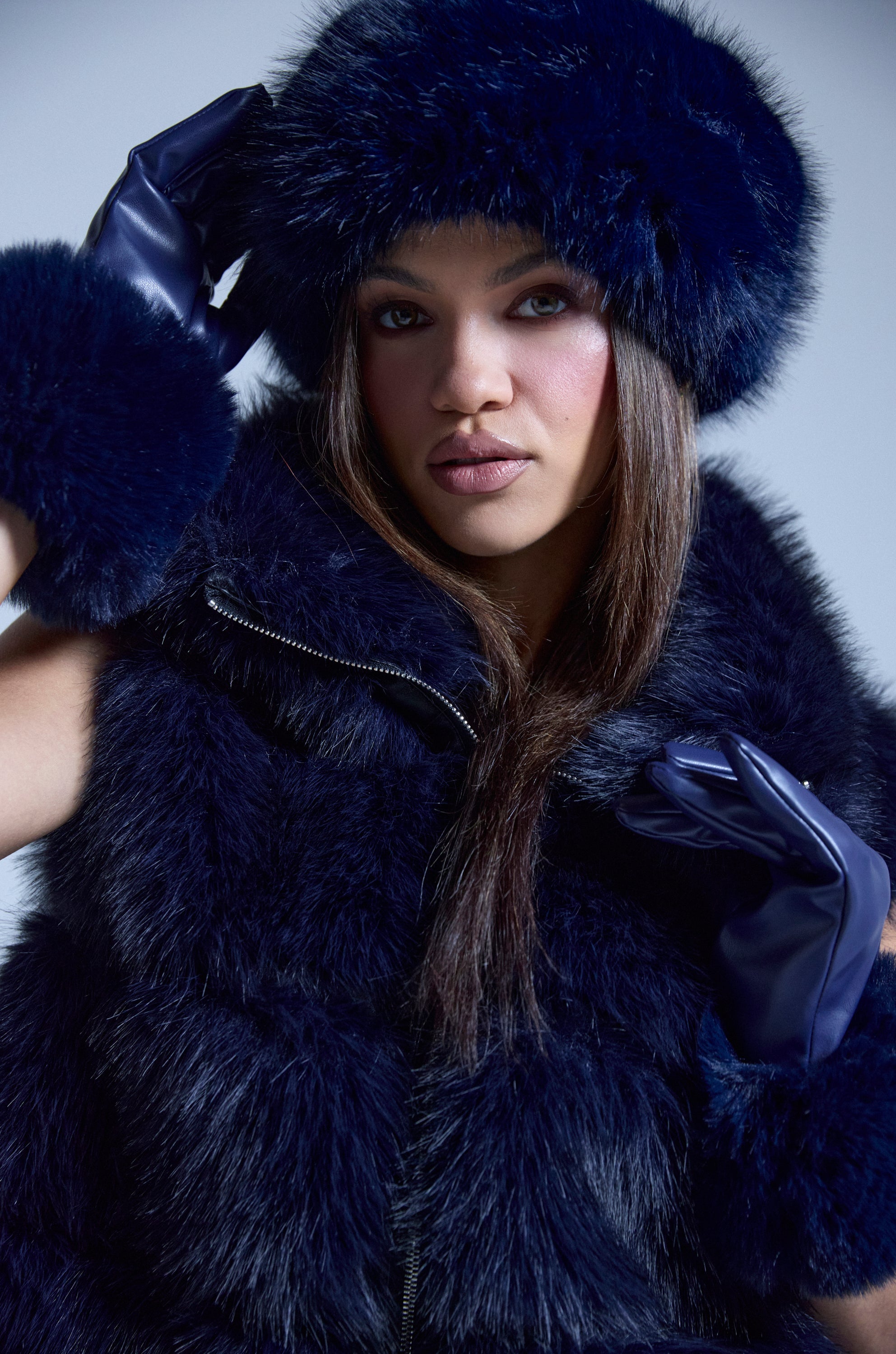 A woman poses confidently against a plain background, wearing the SO CHIC FAUX FUR HEADBAND in dark blue with a matching faux fur vest and gloves, her hands raised near her face as she looks at the camera. 1