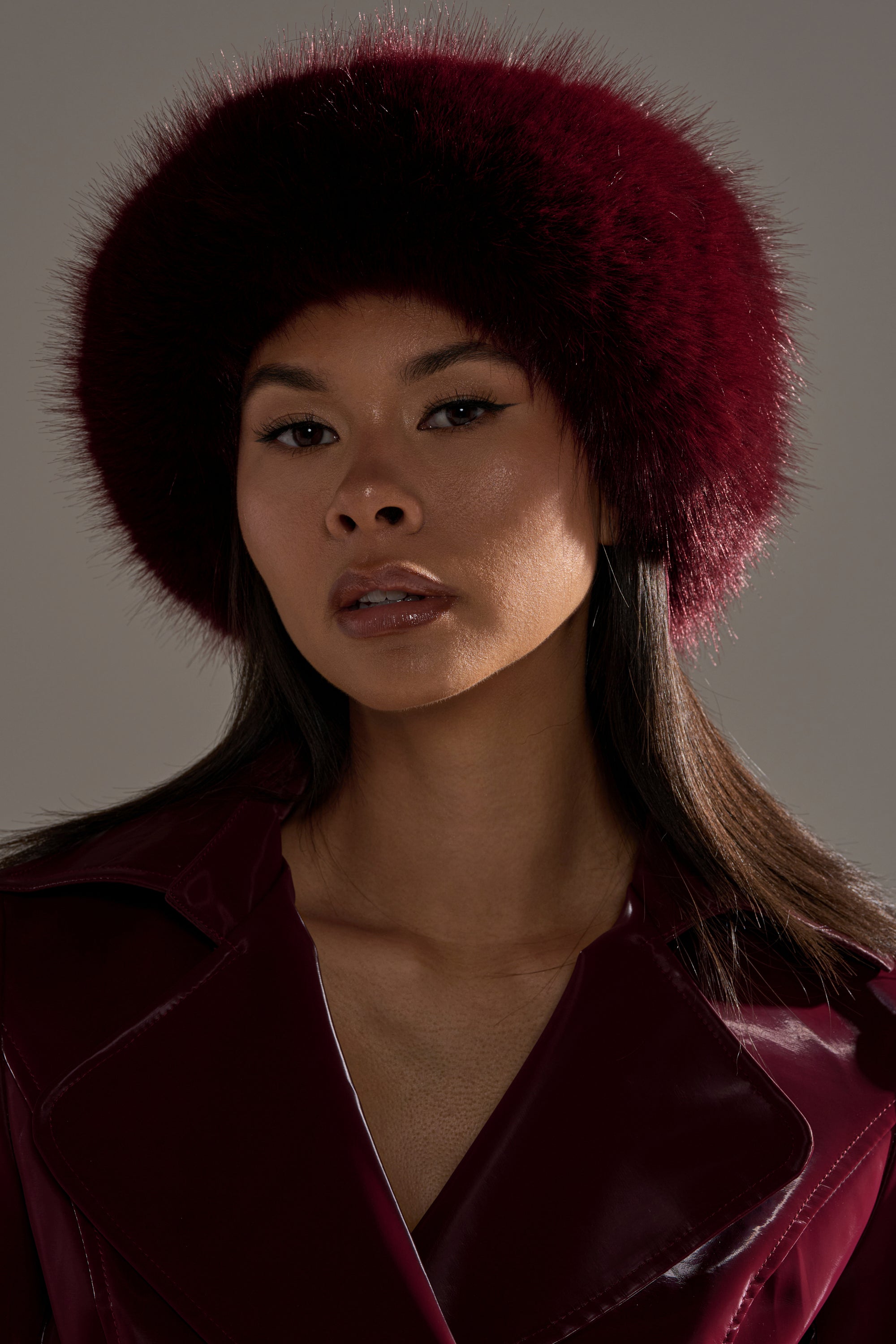 A woman models the ALAIA FAUX FUR HEADBAND, a chic burgundy cold-weather accessory, paired with a glossy dark red coat and set against a neutral background.