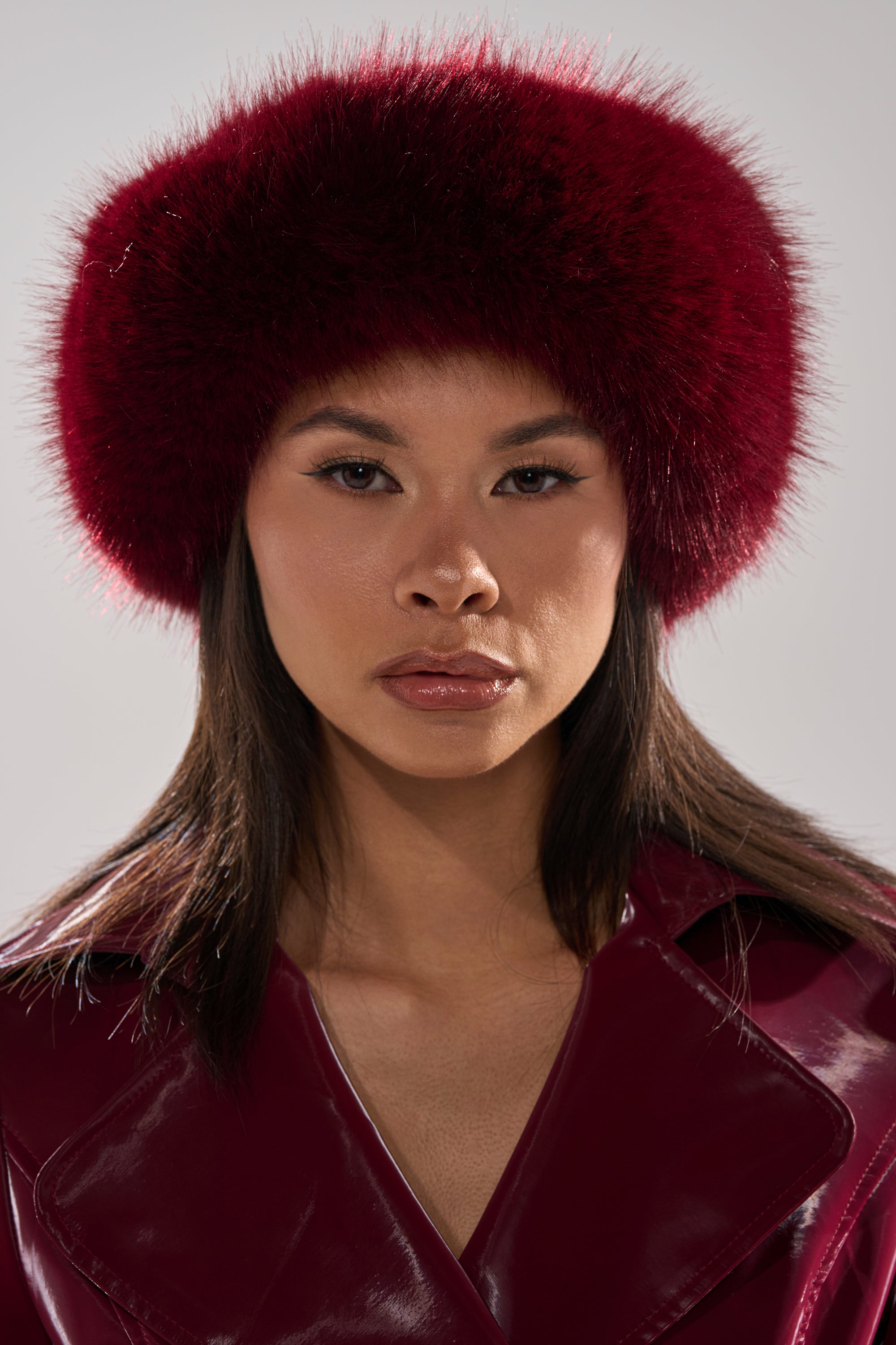 A woman models the ALAIA FAUX FUR HEADBAND in dark red, paired with a matching glossy burgundy coat, looking directly at the camera with a neutral expression against a light gray background.