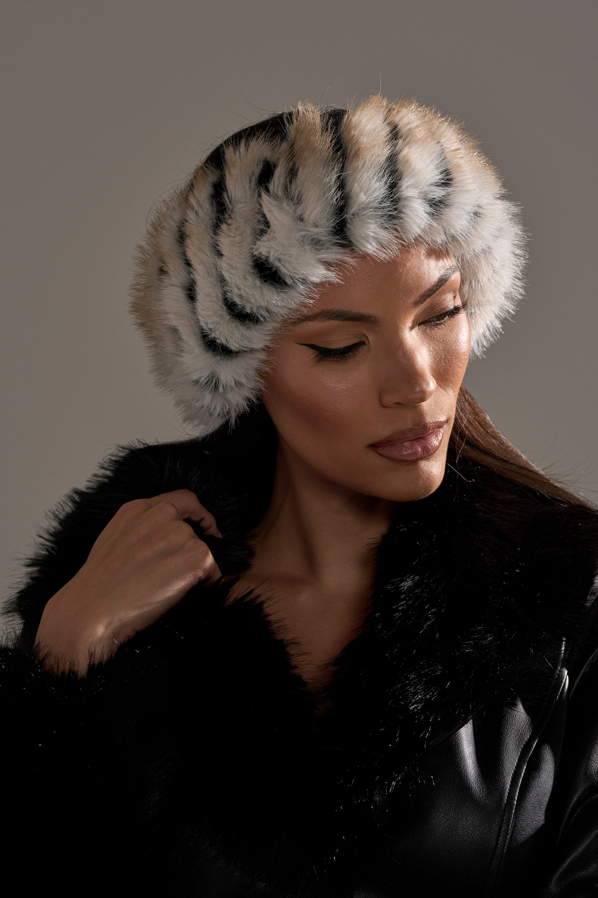 A woman in a black faux fur coat wears the PORTIA FAUX FUR HEADBAND, featuring white and black tiger-like stripes, as she looks down with a soft expression against a neutral background. 0