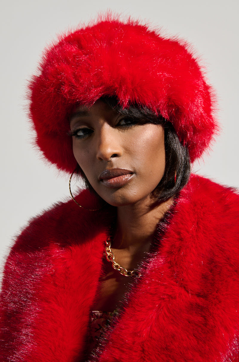 Wearing a statement piece, the individual sports the LOOK AT ME NOW FAUX FUR HEADBAND in a vibrant red hue, complemented by a matching coat. Their long, dark hair flows beneath the headband, while large hoop earrings and a gold necklace add a touch of elegance. They gaze directly into the camera against a simple backdrop. 1