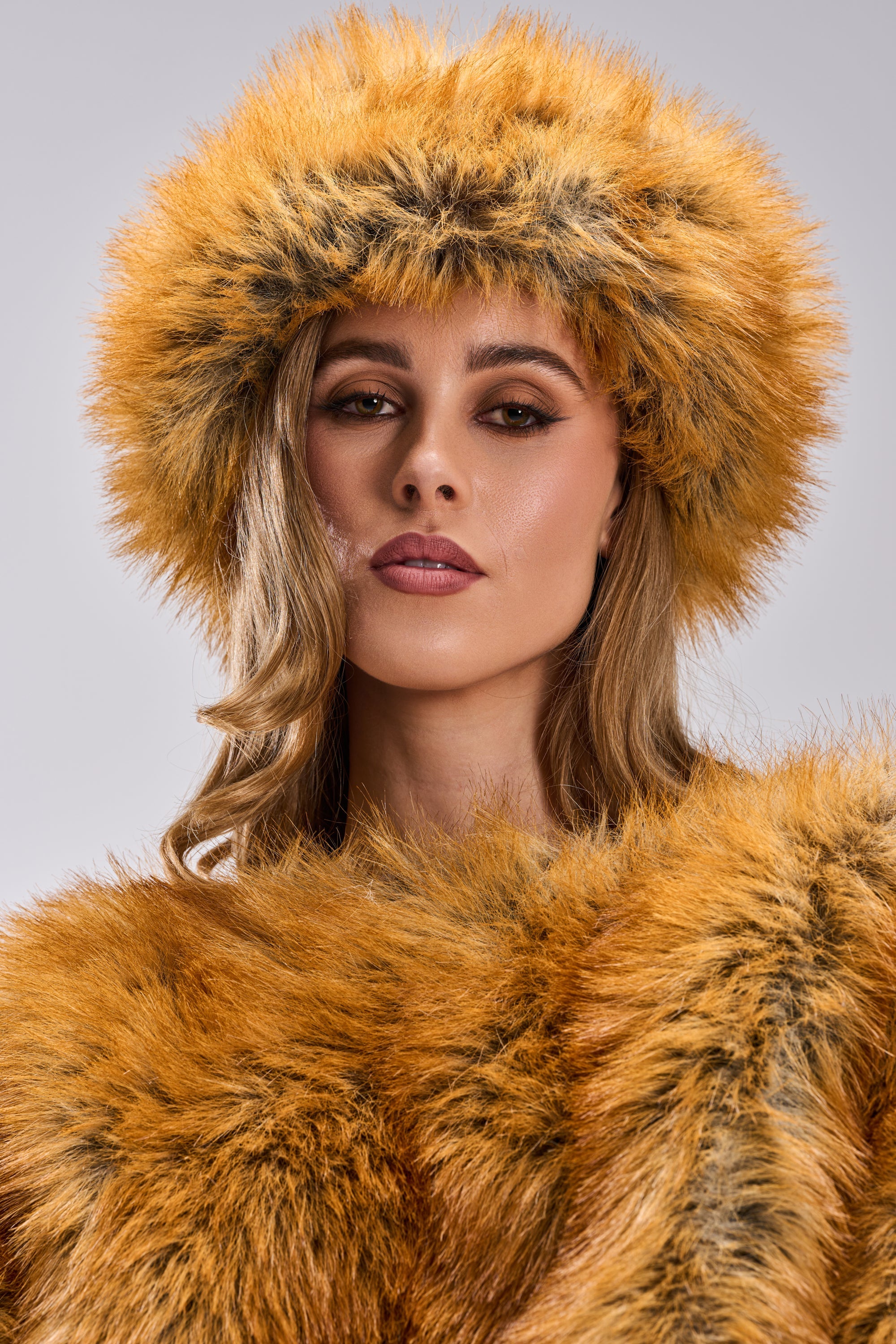 A woman with wavy hair and neutral makeup wears the REALLY LIKE THAT FAUX FUR HEADBAND in orange-brown faux fox fur, confidently facing the camera against a gray background—a chic cold-weather accessory. 3