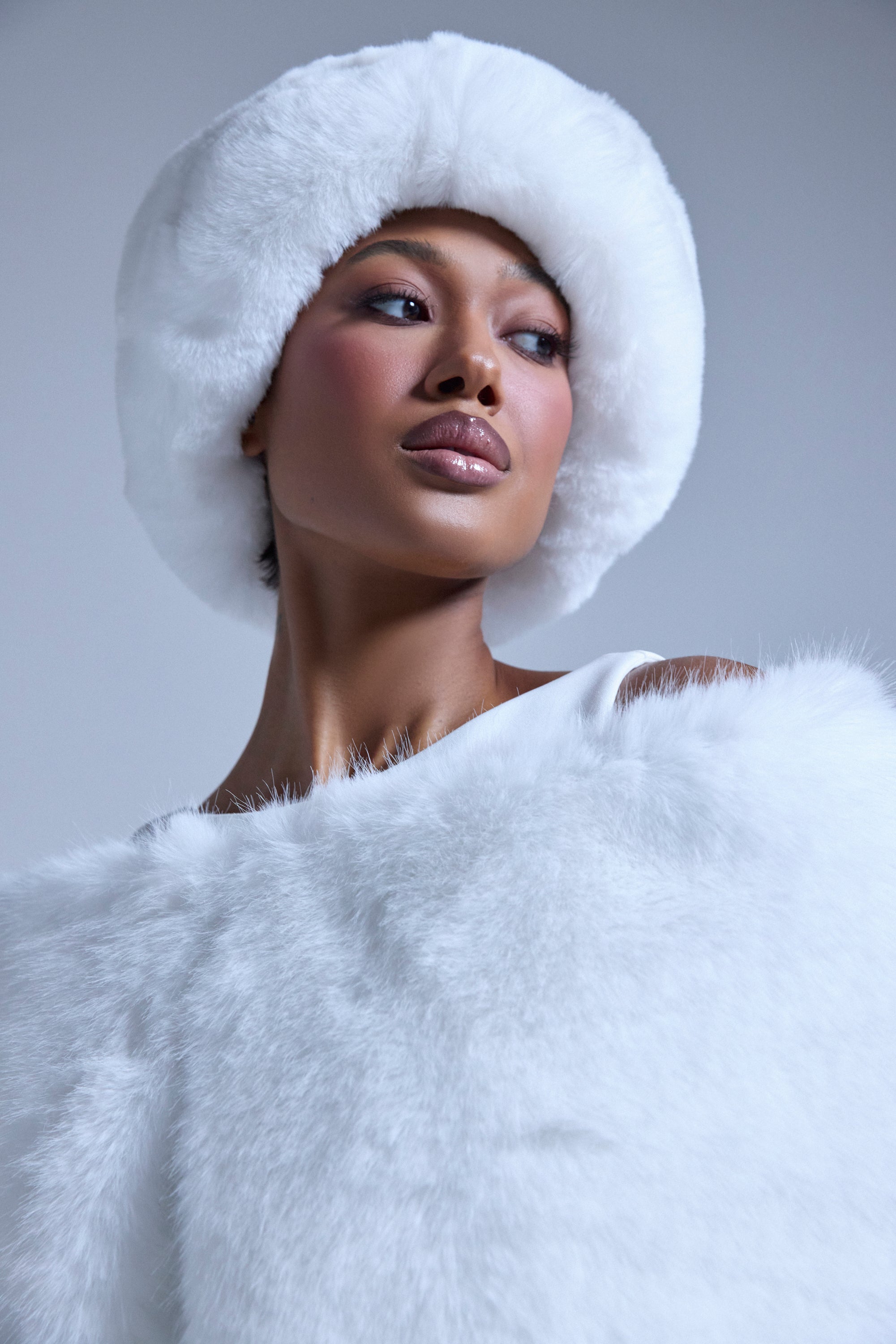 A woman poses confidently against a plain light background, wearing the LUXE JENNY FAUX FUR HEADBAND IN WHITE and a matching off-shoulder faux fur piece.