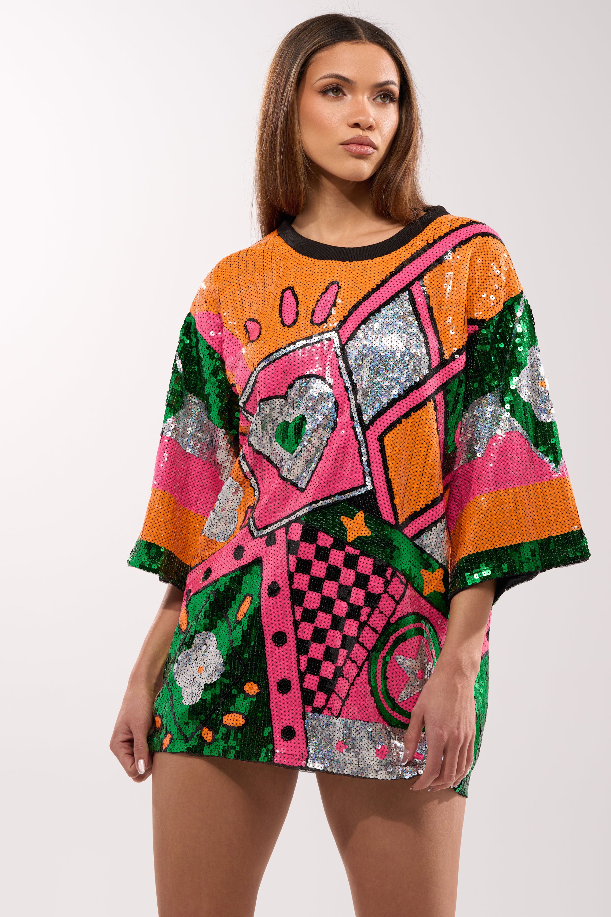 A woman stands against a plain light background wearing the HOUSE OF HEARTS SEQUIN TSHIRT DRESS, a colorful oversized dress with bold geometric patterns, hearts, stars, and floral sequin designs in orange, green, pink, and silver.