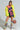 A person poses confidently in a colorful outfit featuring a neon yellow furry jacket, an 80’s inspired HOUSE OF HEARTS SEQUIN TSHIRT DRESS, and matching knee-high boots. They hold a basketball wrapped in rope, showcasing a bold and artistic style.