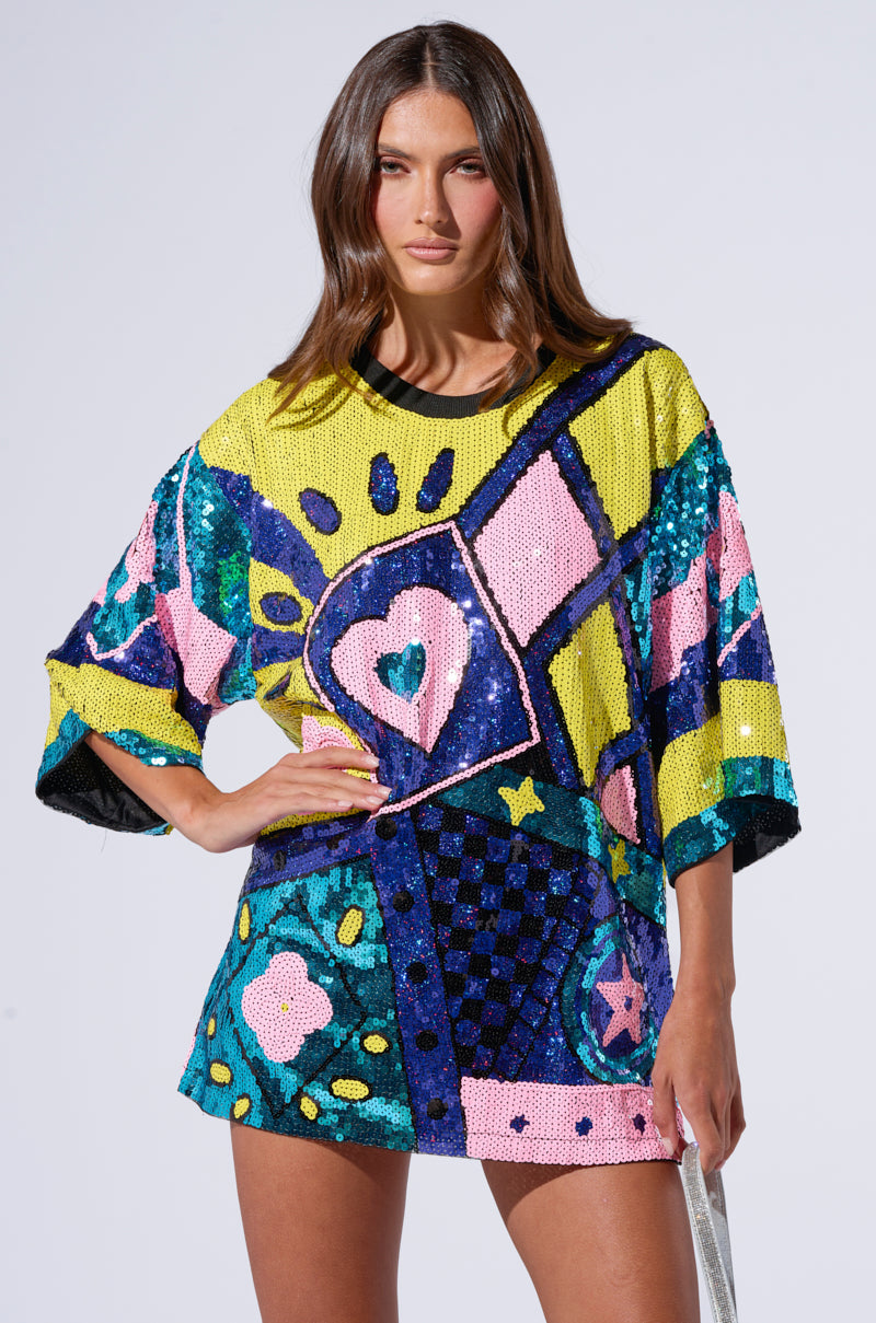 A woman with long brown hair poses with one hand on her hip against a plain backdrop, showcasing the QUEEN OF HEARTS SEQUIN TSHIRT DRESS. The dress is an oversized top adorned with stunning sequin embellishments in pink, blue, yellow, and green, featuring an array of bright patterns and shapes.