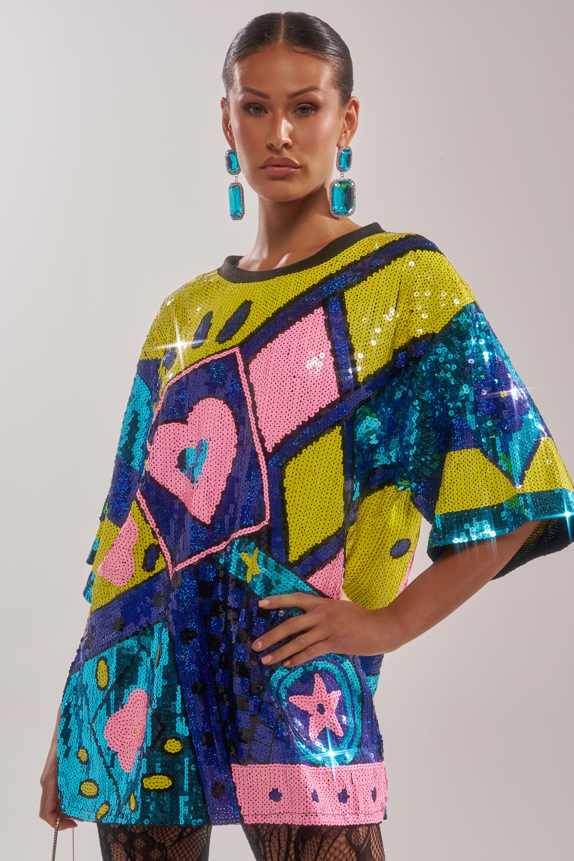 A woman models the QUEEN OF HEARTS SEQUIN TSHIRT DRESS, a bold, colorful oversized dress embellished with sequins, geometric shapes, hearts, and stars. She pairs it with blue earrings and slicked-back hair for a striking look.