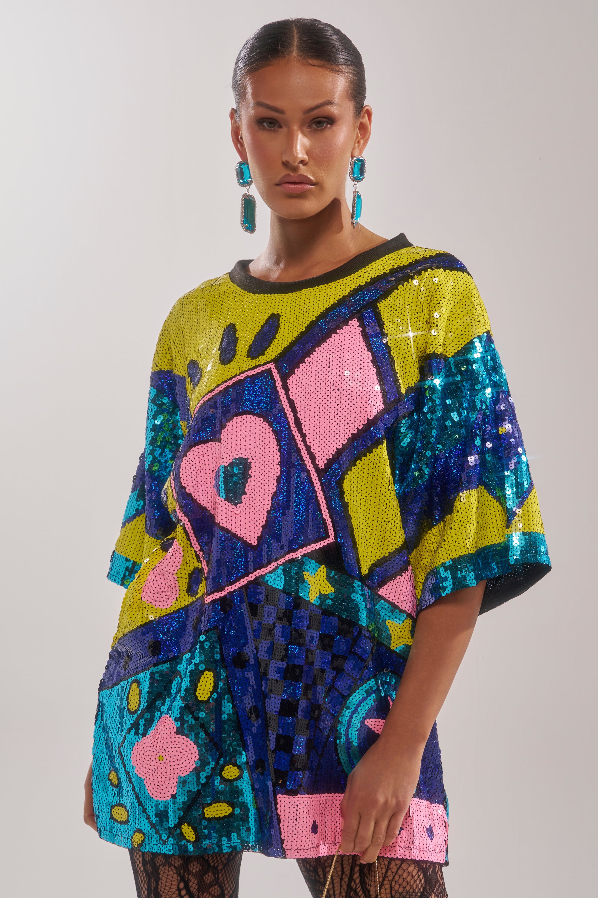 A woman stands against a plain background, wearing the QUEEN OF HEARTS SEQUIN TSHIRT DRESS—an oversized dress with colorful geometric and abstract sequin patterns—paired with statement blue earrings and patterned tights.