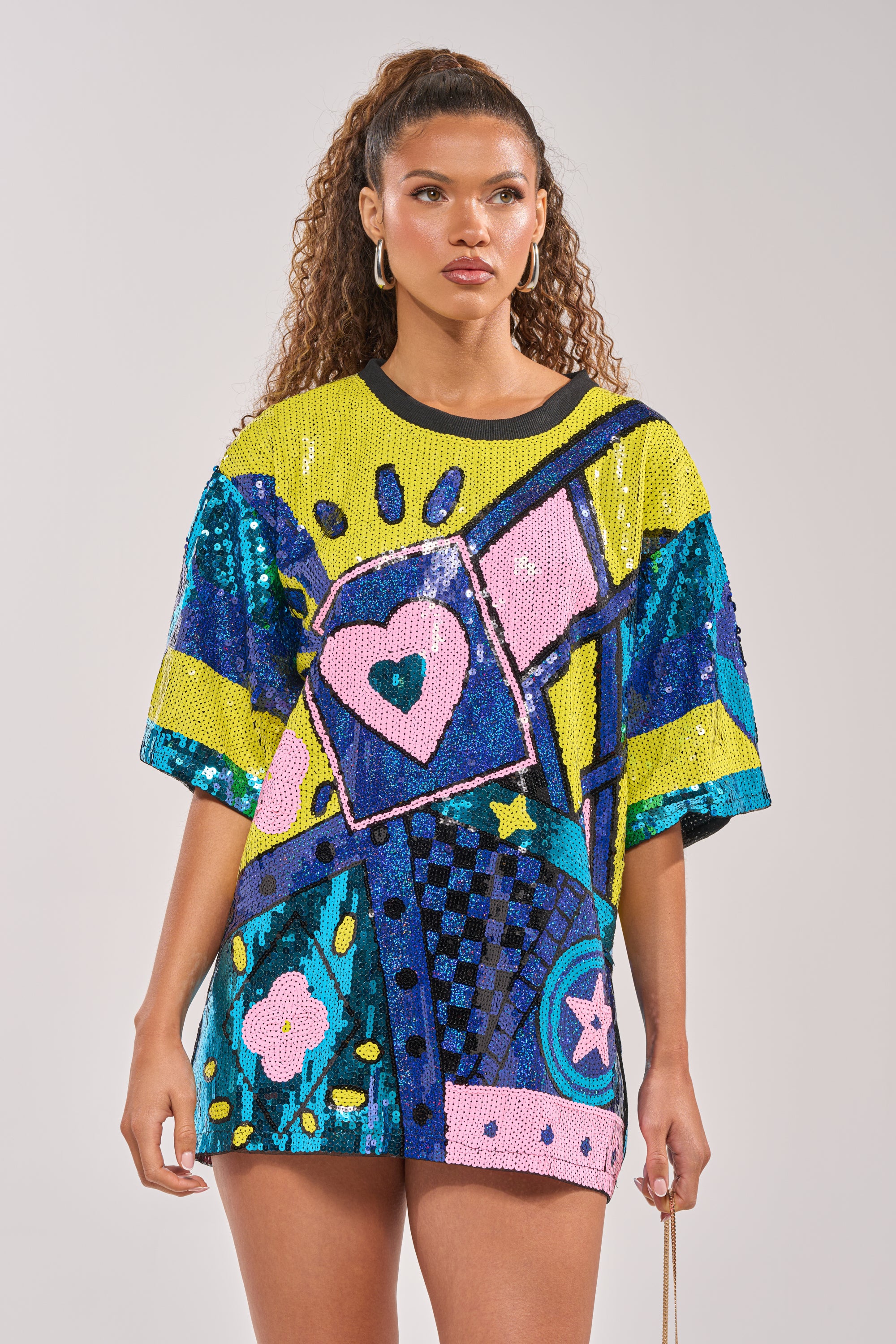 A woman wears the QUEEN OF HEARTS SEQUIN TSHIRT DRESS, featuring vibrant abstract hearts, flowers, and geometric patterns in blue, yellow, pink, and green with multicolor block paneling. Her curly hair is styled in a high ponytail with large hoop earrings. 1