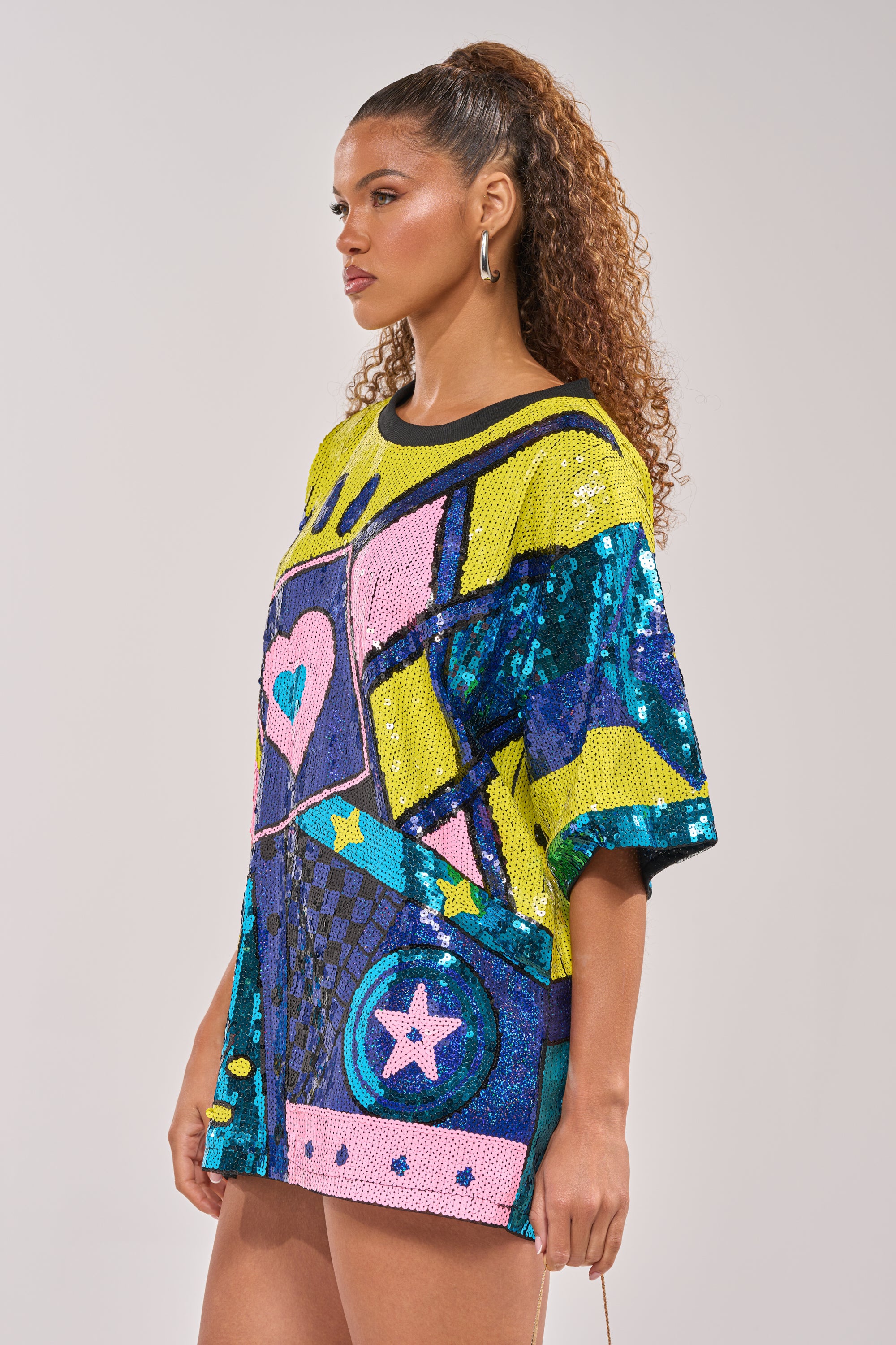 A woman with curly hair in a high ponytail wears the QUEEN OF HEARTS SEQUIN TSHIRT DRESS featuring bold geometric shapes, stars, and a heart. She stands sideways with a neutral expression against a plain background. 2