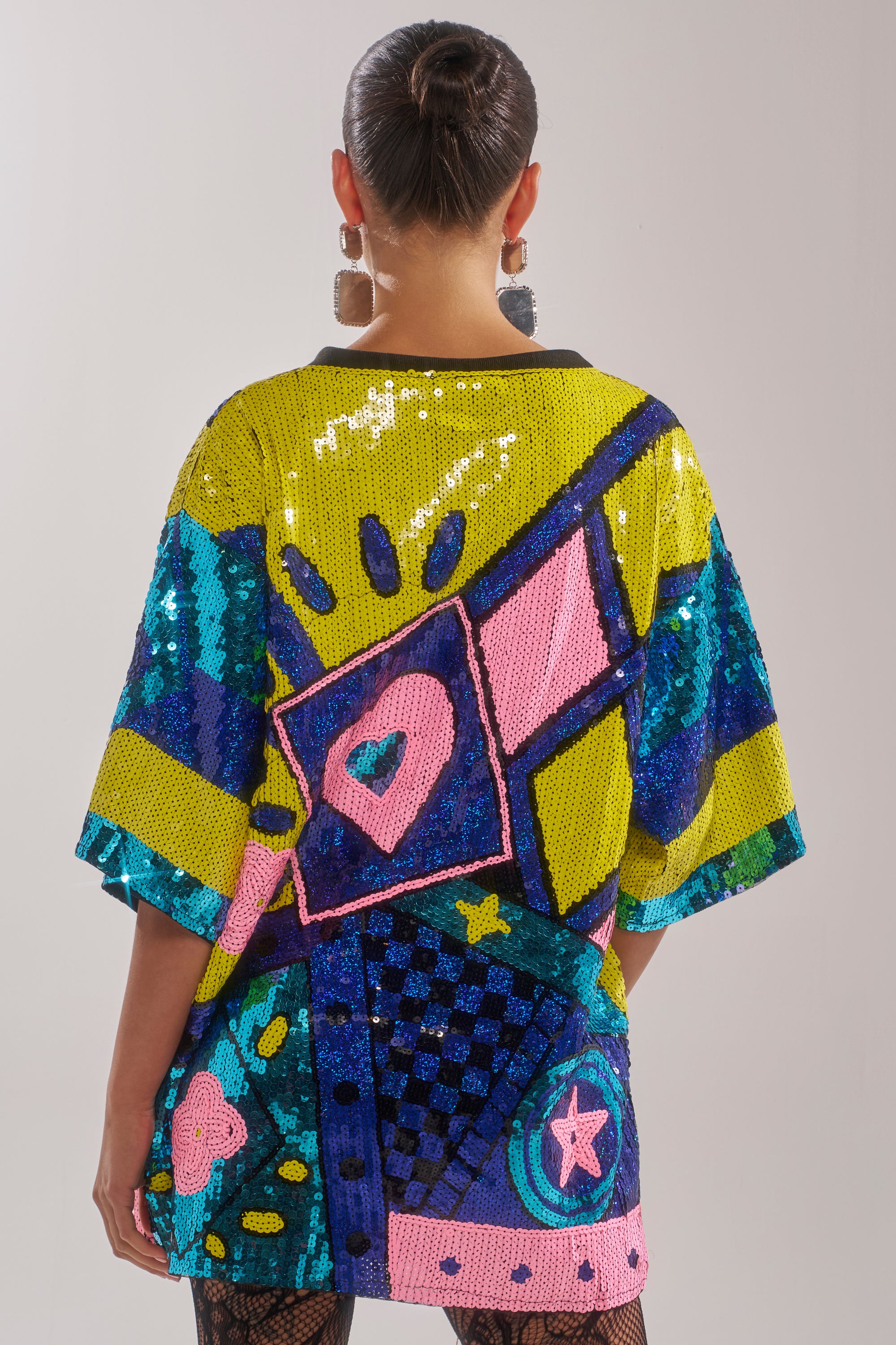 A person with dark hair in a bun is seen from the back wearing the QUEEN OF HEARTS SEQUIN TSHIRT DRESS, an oversized piece adorned with colorful geometric and abstract heart and star patterns in yellow, blue, pink, and green.