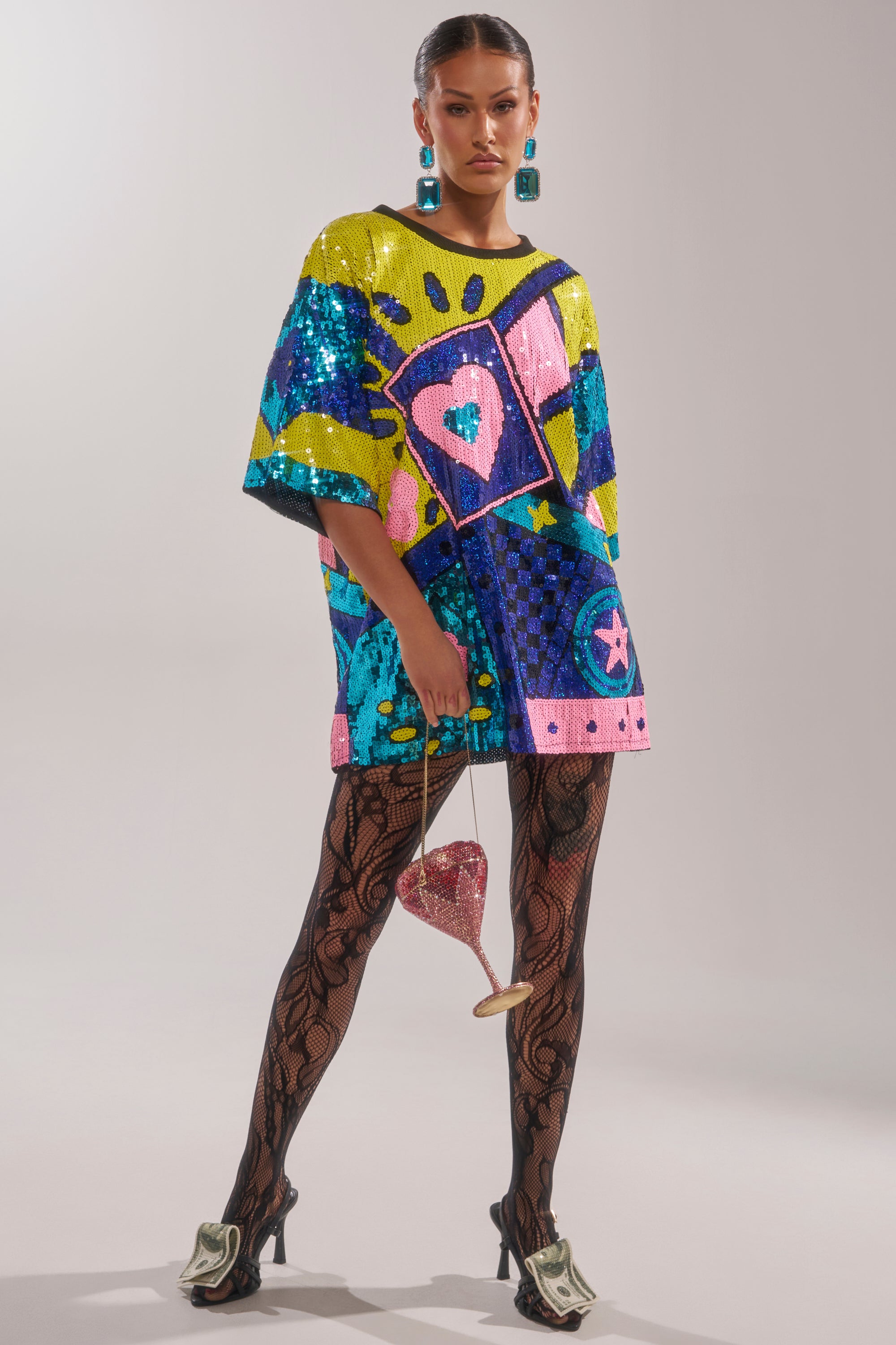 A woman confidently wears the QUEEN OF HEARTS SEQUIN TSHIRT DRESS, pairing it with large blue earrings, black lace-patterned tights, heels decorated with dollar bills, and carries a small pink heart-shaped purse.