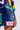 A close-up image of a person wearing the QUEEN OF HEARTS SEQUIN TSHIRT DRESS, showcasing its vibrant, multi-colored geometric patchwork design with stars, checkered patterns, and various shapes in shades of blue, pink, yellow, green, and purple. The person's hand is resting by their side.