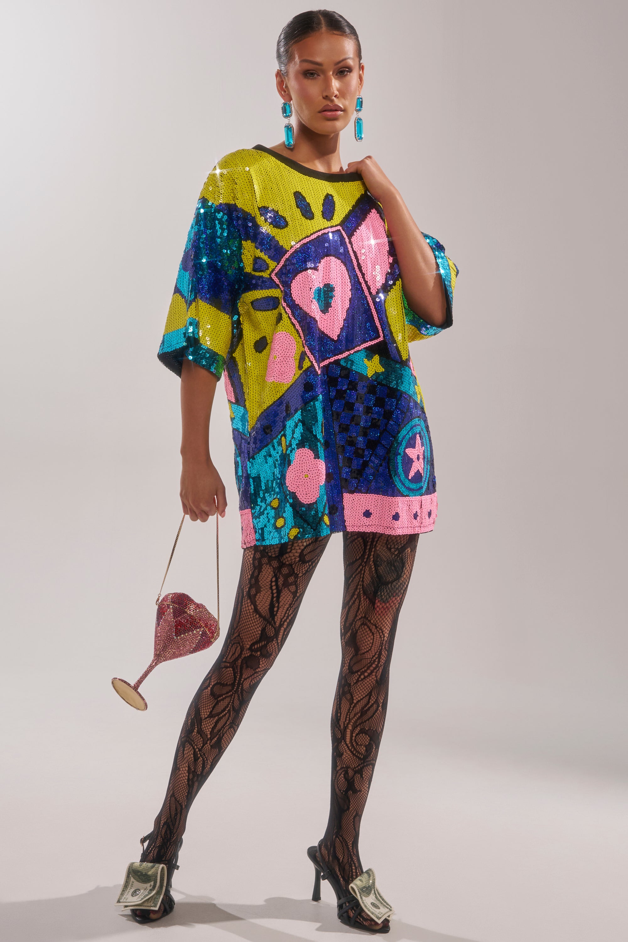 A woman poses in the QUEEN OF HEARTS SEQUIN TSHIRT DRESS, featuring vibrant sequins and abstract designs, paired with black tights, dollar bill high heels, blue earrings, and a sparkling wine glass-shaped purse—a true statement look.