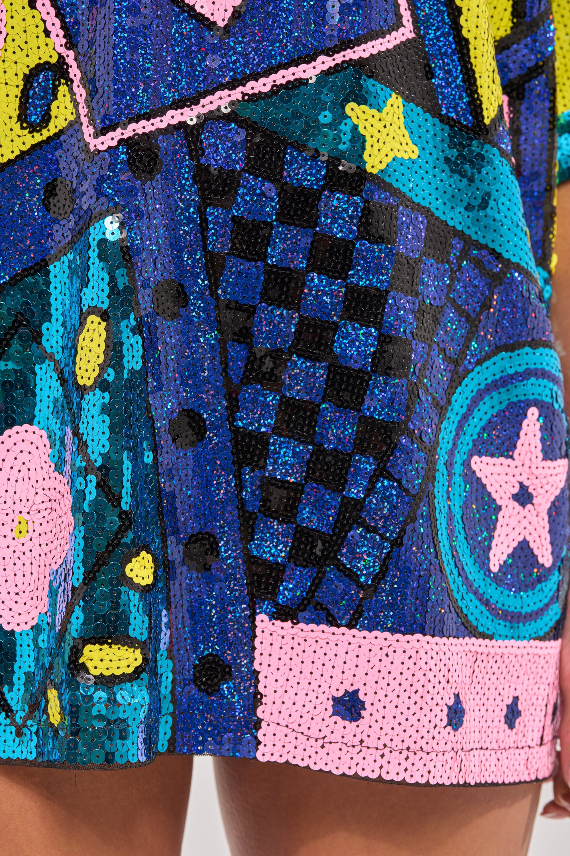 Close-up of the QUEEN OF HEARTS SEQUIN TSHIRT DRESS, featuring colorful star, check, and abstract sequin patterns in blue, pink, yellow, and black. The hem and part of a person's leg are visible at the bottom. 7