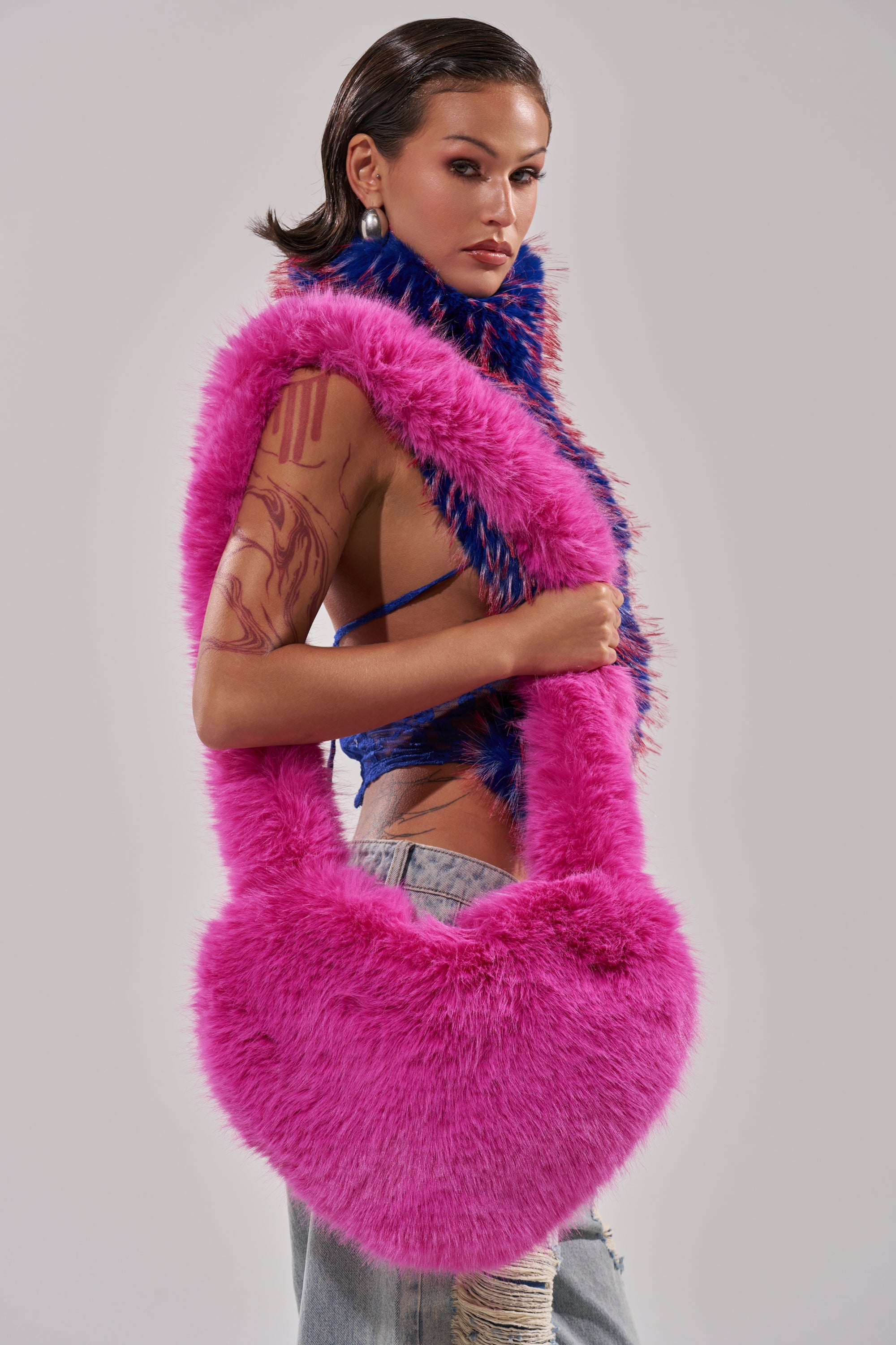 A person with slicked-back hair, pearl earrings, a blue textured top, distressed jeans, and a visible upper arm tattoo poses holding the LANA FAUX FUR HEART BAG IN PINK over their shoulder. 1