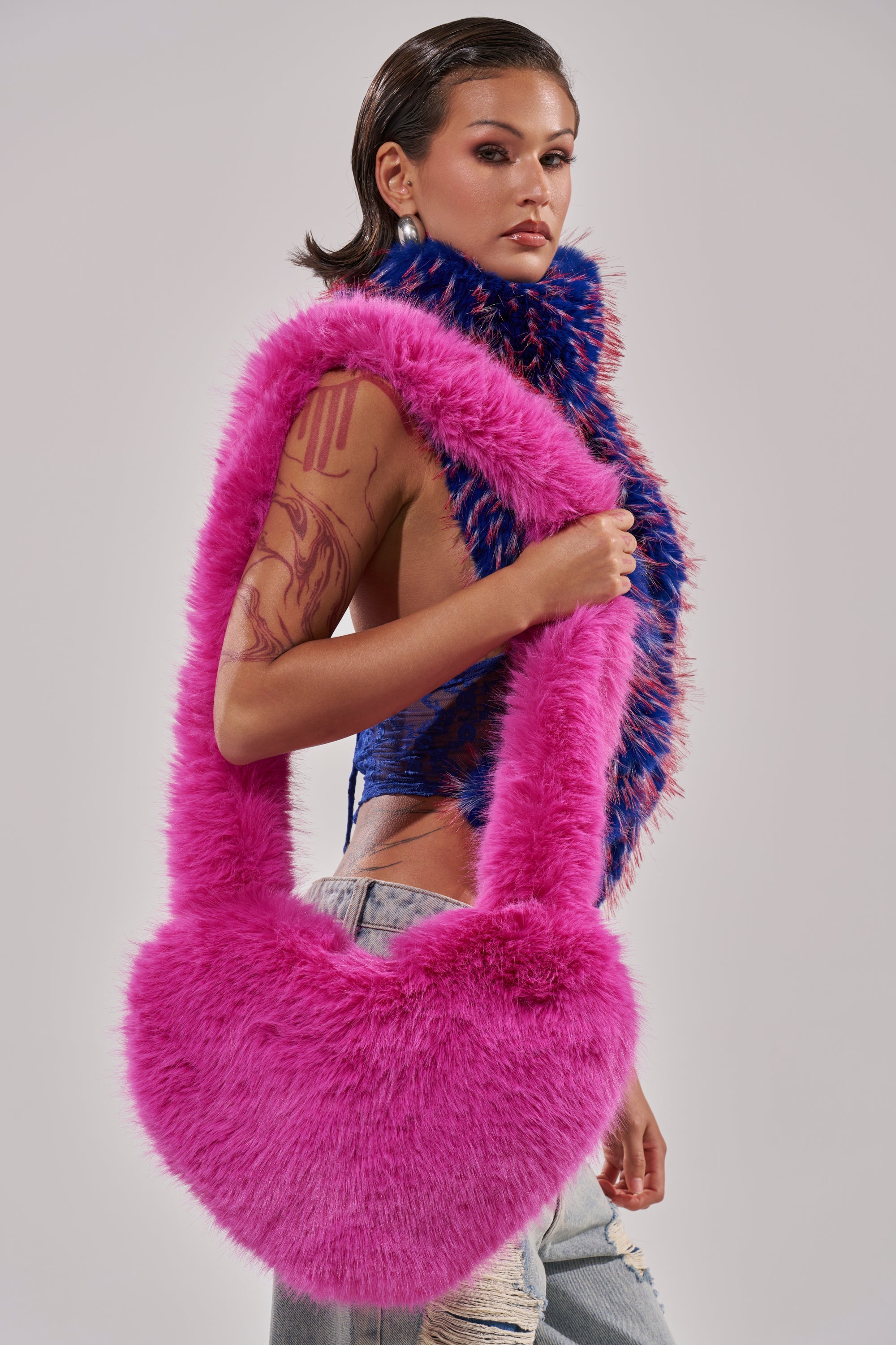 A woman with slicked-back hair and a tattooed arm poses in a blue top, ripped jeans, and a colorful furry scarf. She carries the LANA FAUX FUR HEART BAG IN PINK over her shoulder. 4