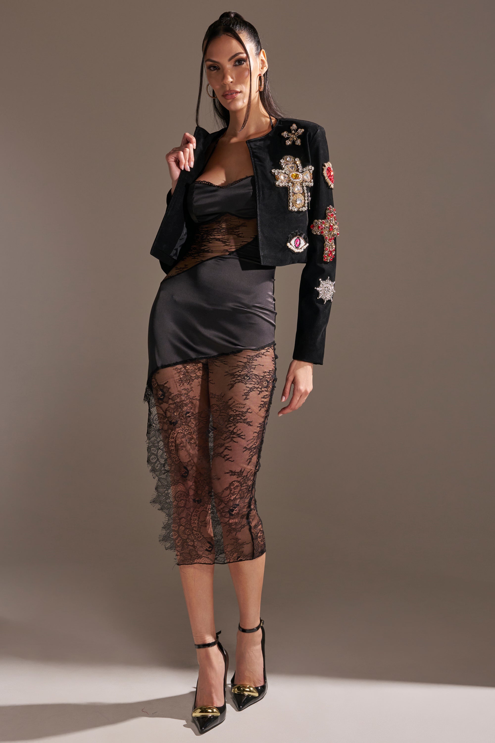 A woman poses in a sheer black lace dress, black mini skirt, and the COUNT YOUR BLESSINGS EMBELLISHED BLAZER, with black heels featuring gold pointed toes, against a plain gray background. 9