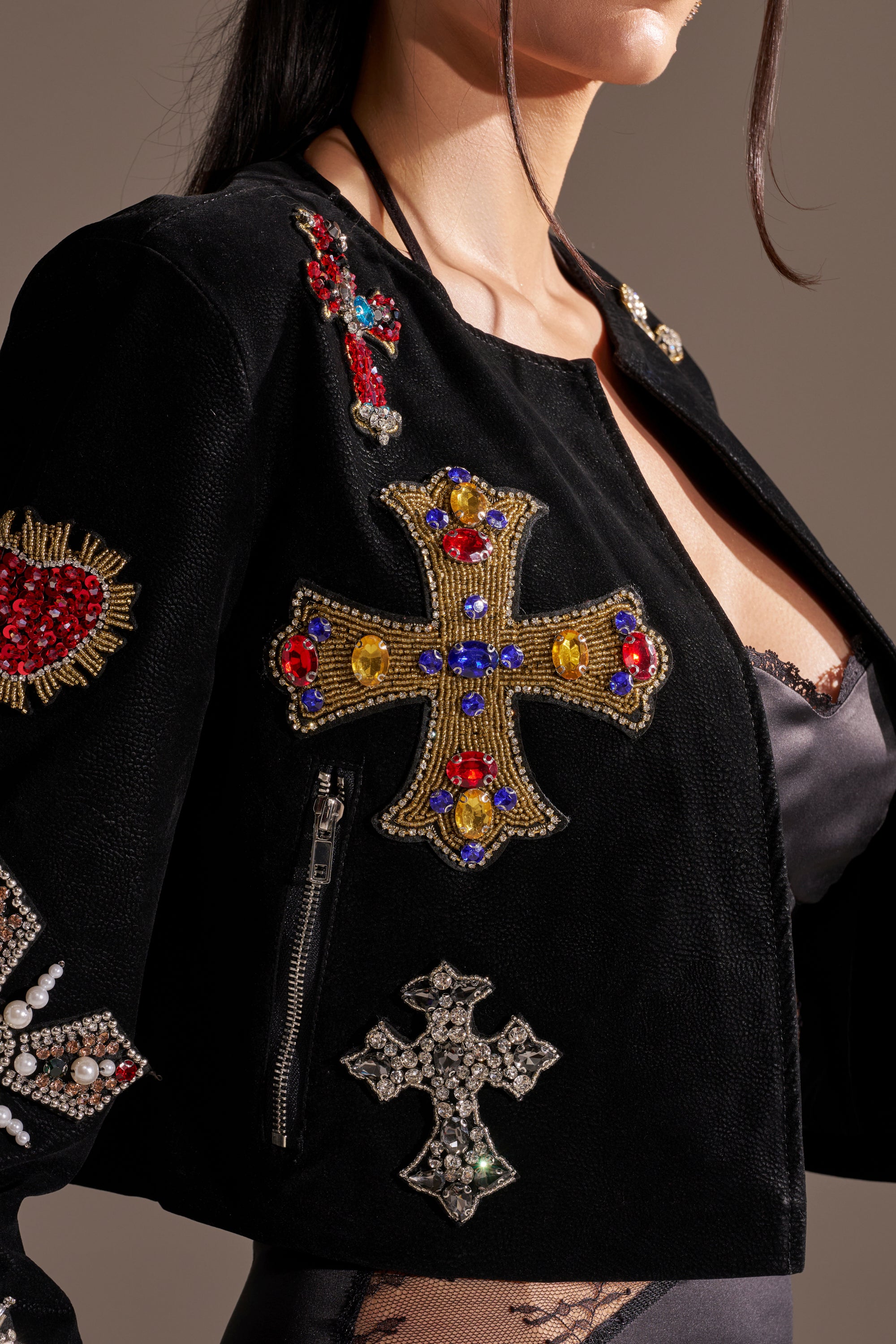 A woman wears the COUNT YOUR BLESSINGS EMBELLISHED BLAZER, detailed with colorful beaded cross and heart motifs, over a black satin top. The image focuses on her upper torso and the blazer’s intricate embellishments. 10