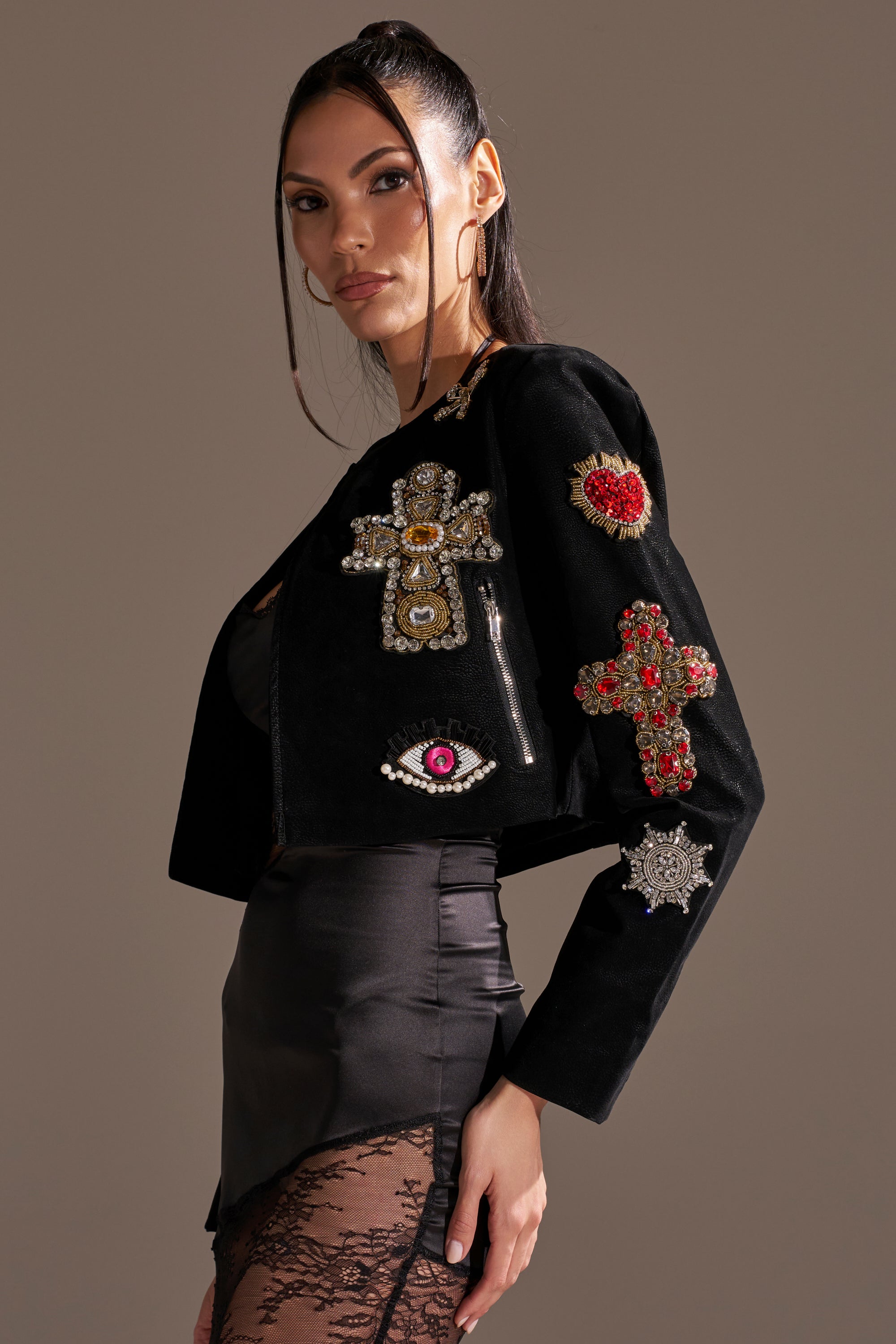 A woman poses in profile wearing the COUNT YOUR BLESSINGS EMBELLISHED BLAZER, featuring black fabric with cross, eye, and heart designs. She pairs it with a black satin and lace skirt and styles her hair in a sleek ponytail. 5
