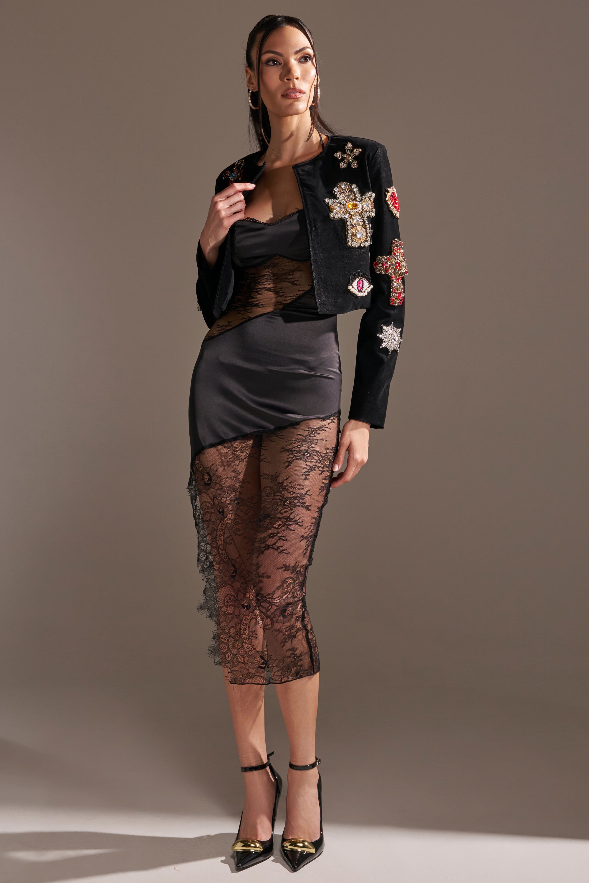 A woman poses confidently in the COUNT YOUR BLESSINGS EMBELLISHED BLAZER, a cropped faux leather jacket with embroidered patches, over a fitted black dress with sheer lace panels against a neutral background. 7
