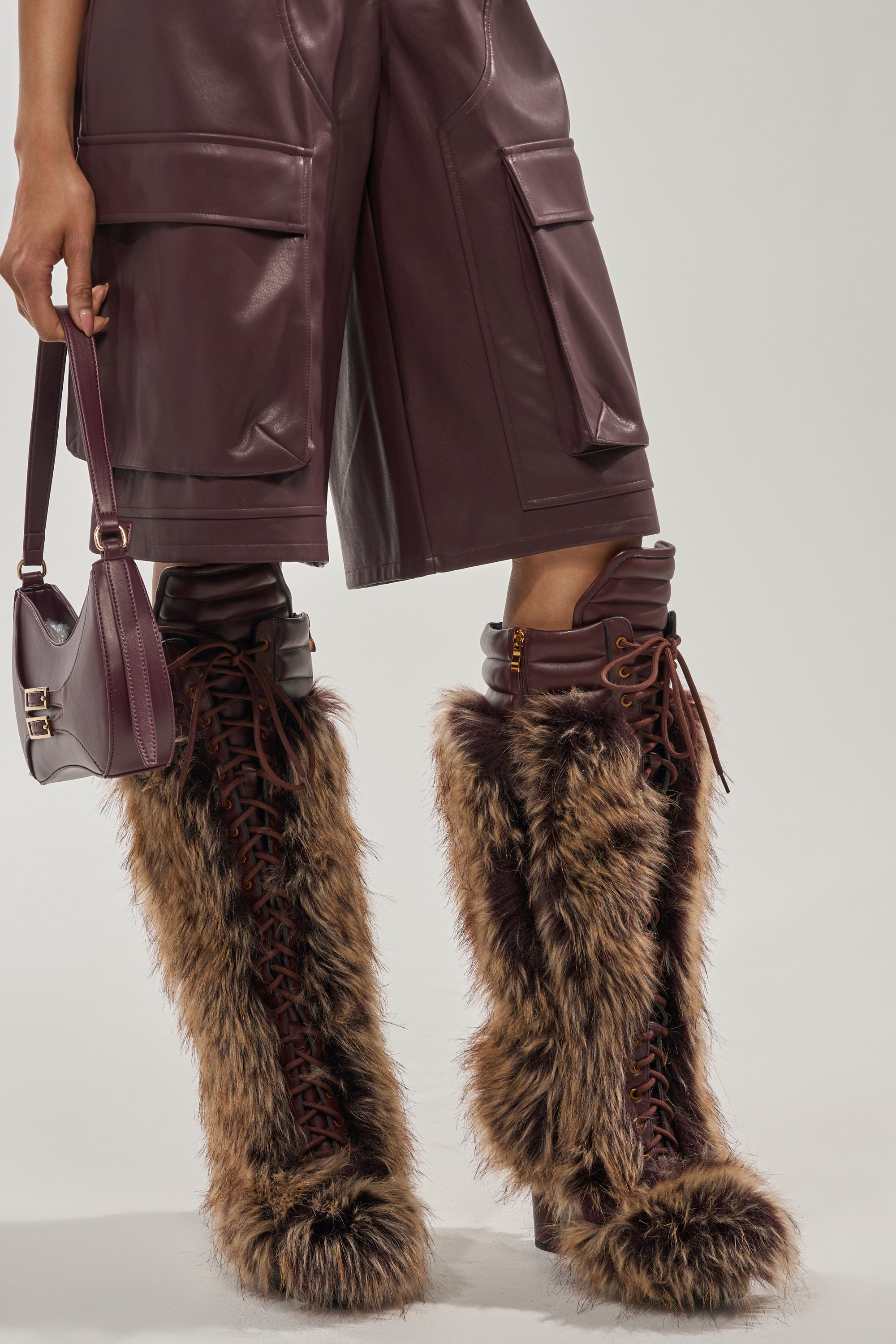 Wearing knee-length burgundy leather cargo shorts and a matching small bag, a person stands against a plain background in AZALEA WANG HEARTBEAT BROWN MULTI BLOCK FUR BOOTS—stylish knee-highs ideal for winter. 6