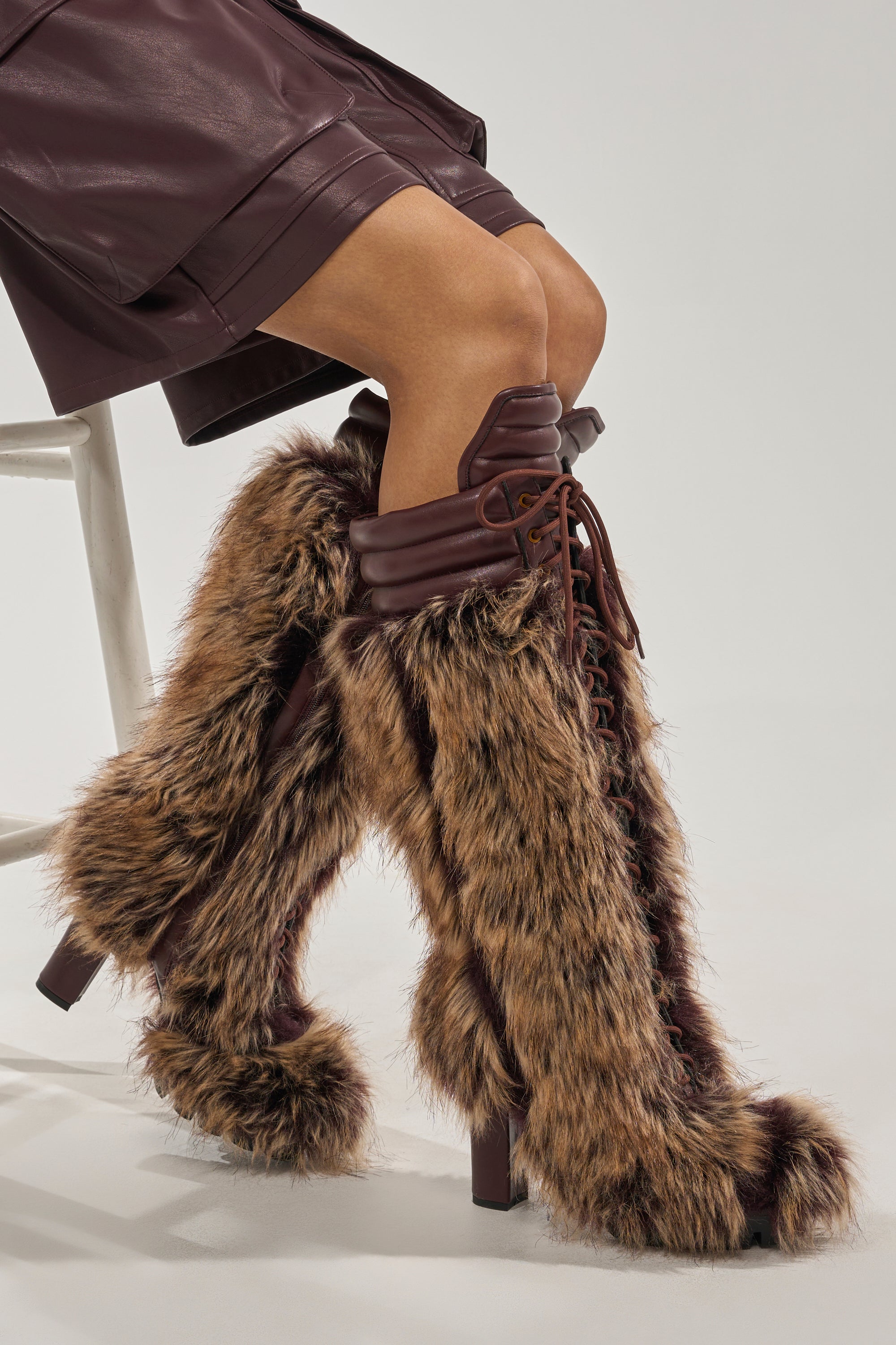 A person seated on a white stool wears a dark brown leather skirt and the AZALEA WANG HEARTBEAT BROWN MULTI BLOCK FUR BOOT—stylish, knee-high winter boots with faux fur and leather accents. 7