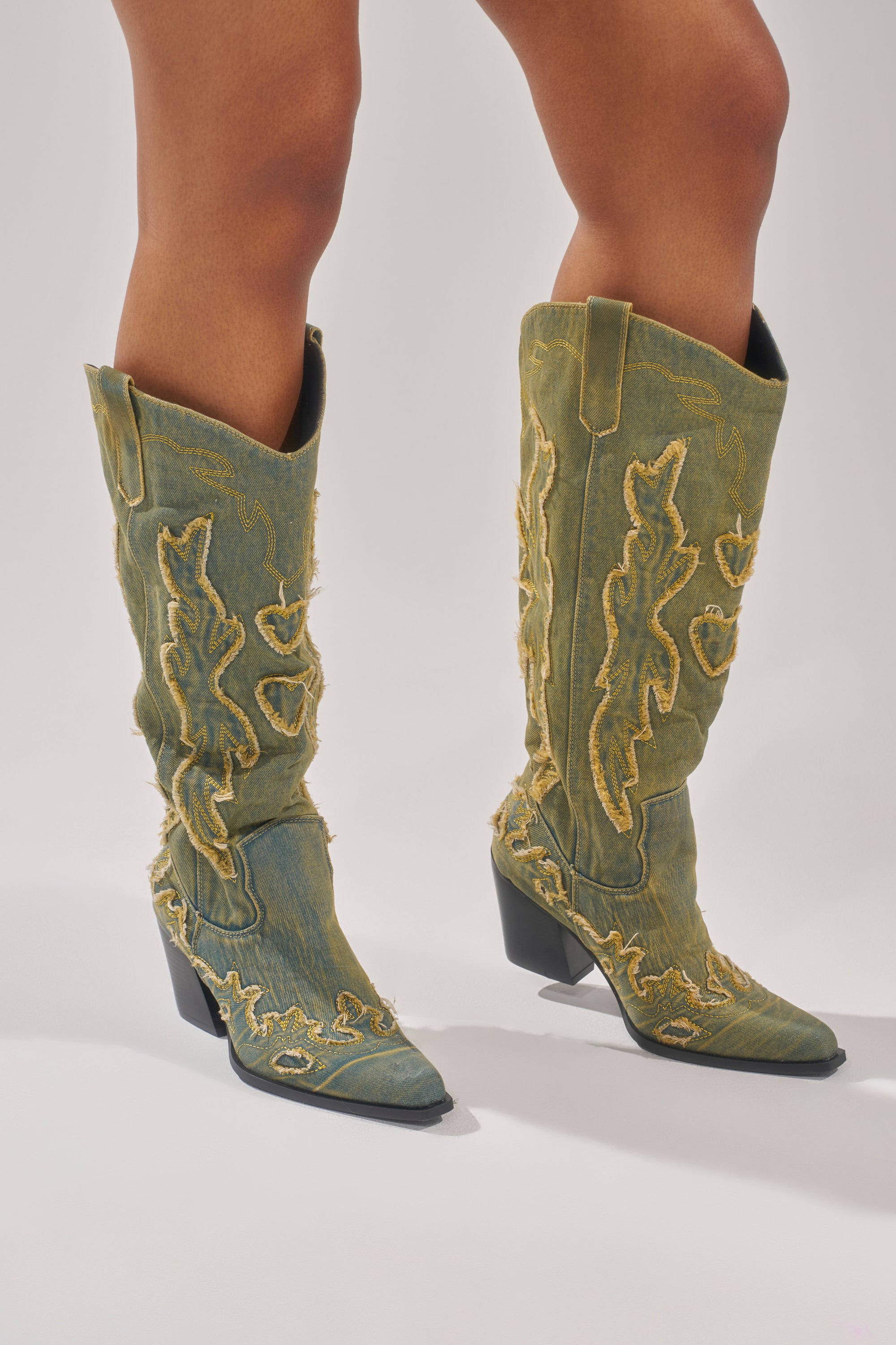 A person wears the AZALEA WANG HEINRICH DENIM WESTERN LOW HEEL BOOT—knee-high, pointed-toe green cowboy boots with yellow frayed stitching—standing on a white background.