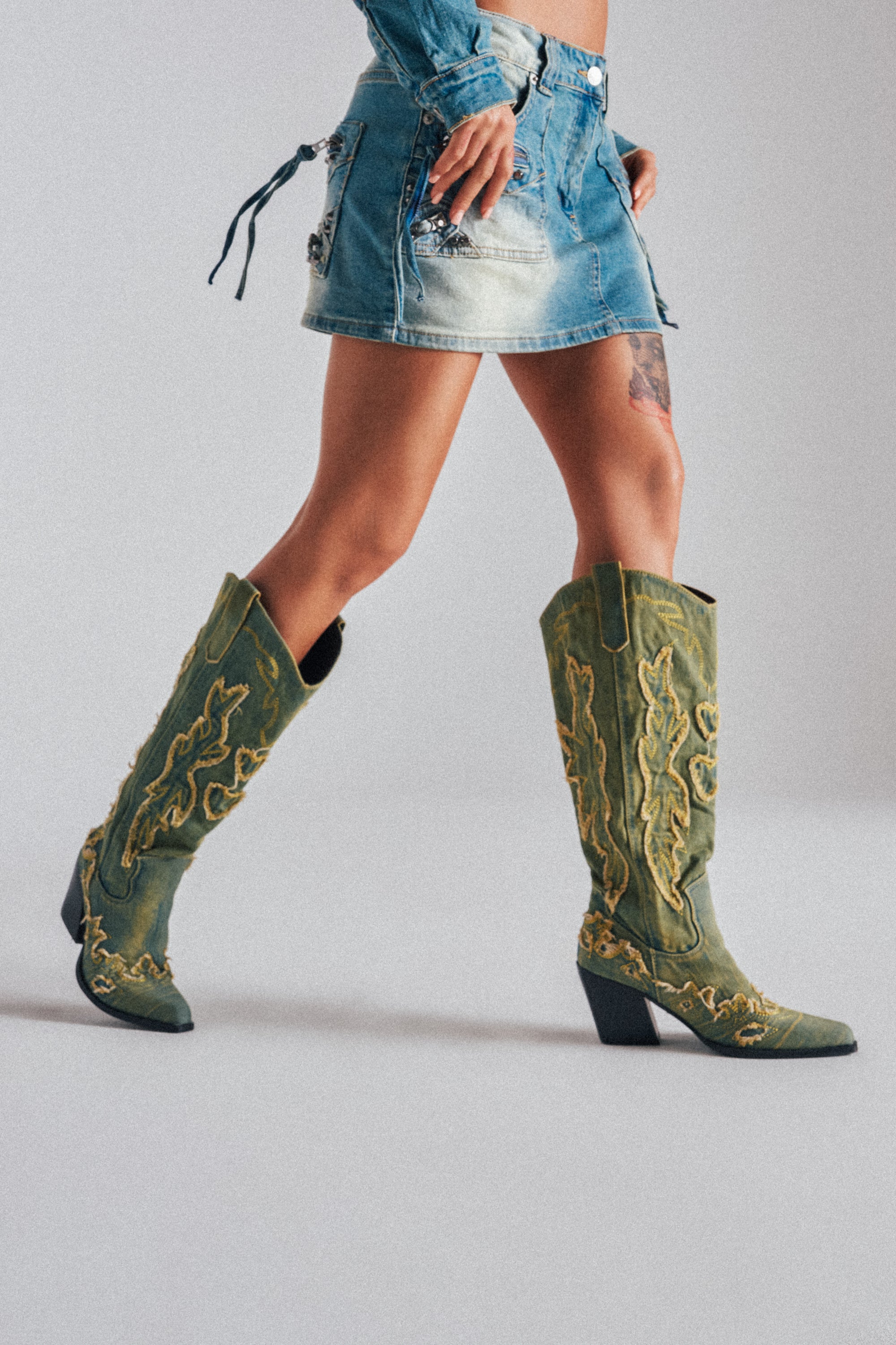 A person wears a short denim skirt and AZALEA WANG HEINRICH DENIM WESTERN LOW HEEL BOOTS, featuring striking green color with yellow embroidery, walking against a plain light background.