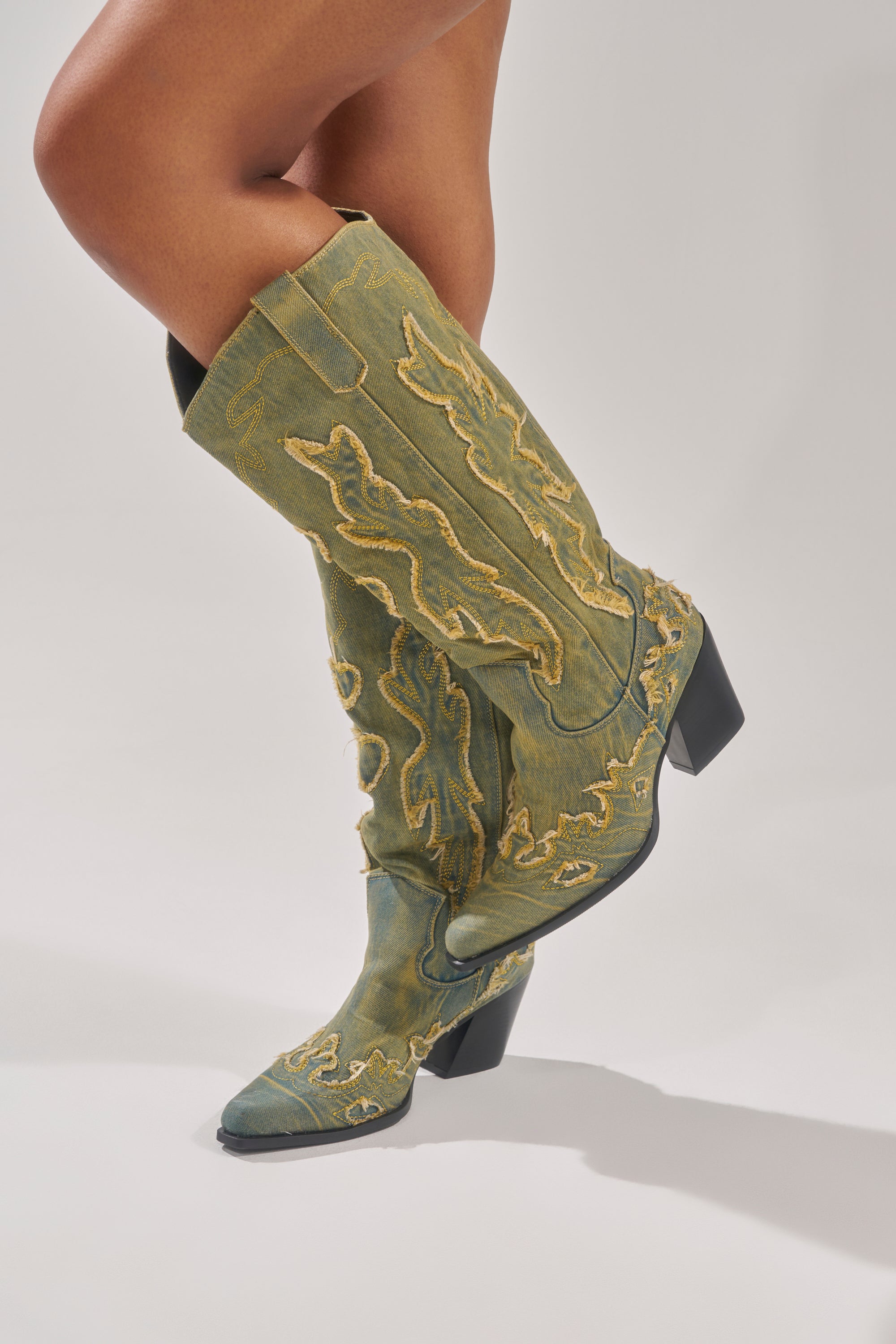 A person stands with one leg crossed over the other in AZALEA WANG HEINRICH DENIM WESTERN LOW HEEL BOOTS—knee-high green denim with yellow stitching and a black heel—against a plain background.