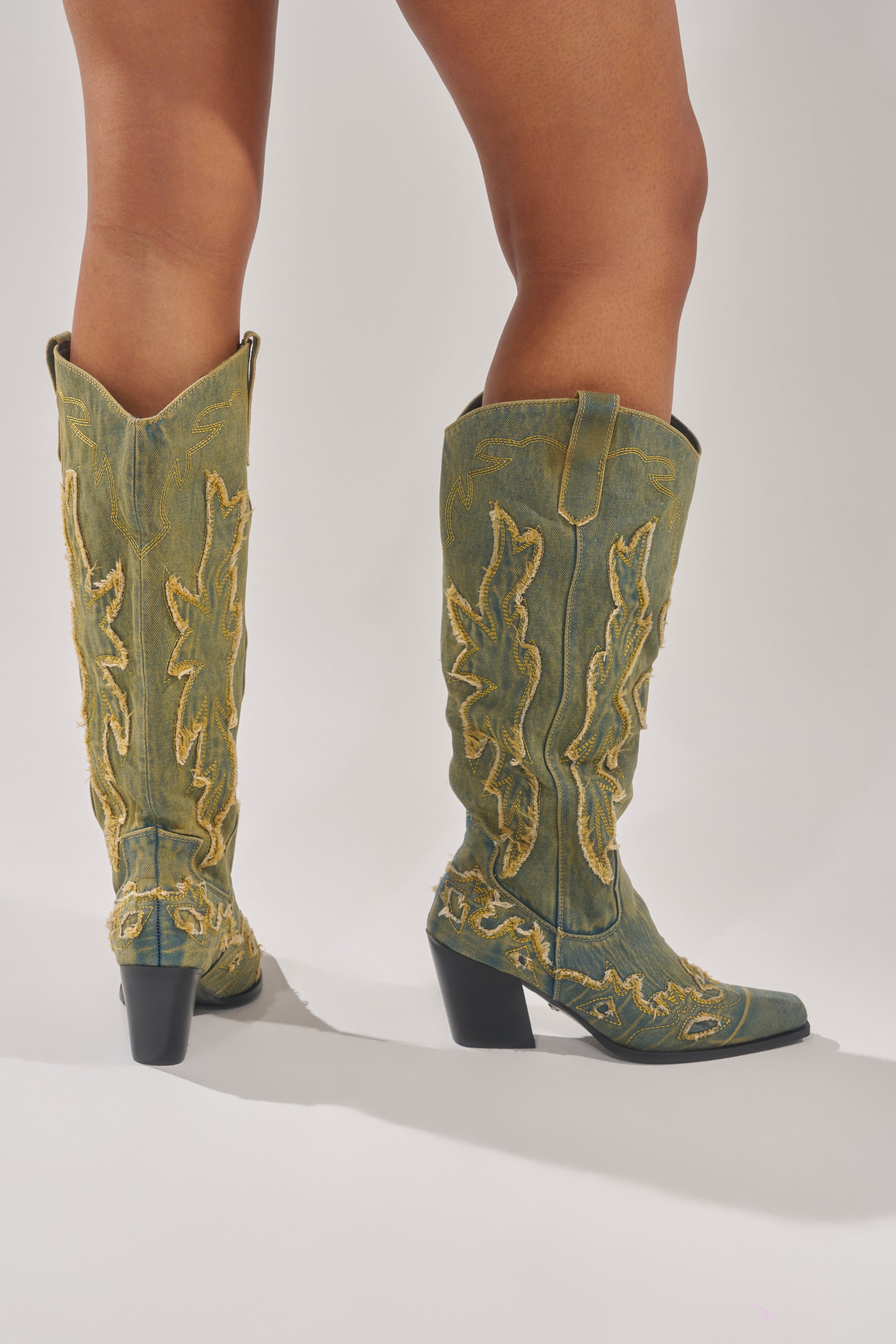 A person stands from lower thigh down, wearing AZALEA WANG HEINRICH DENIM WESTERN LOW HEEL BOOTS featuring greenish denim, yellow embroidery, and chunky black heels against a light background.