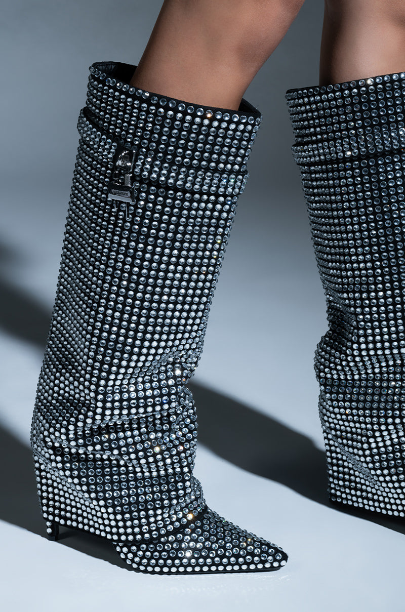 A close-up of an individual wearing the AZALEA WANG HEMLOCK SILVER RHINESTONE STILETTO BOOT, adorned with numerous shimmering silver gems. These knee-high boots showcase a slouched design and pointed toes, set against a simple, neutral grey background.