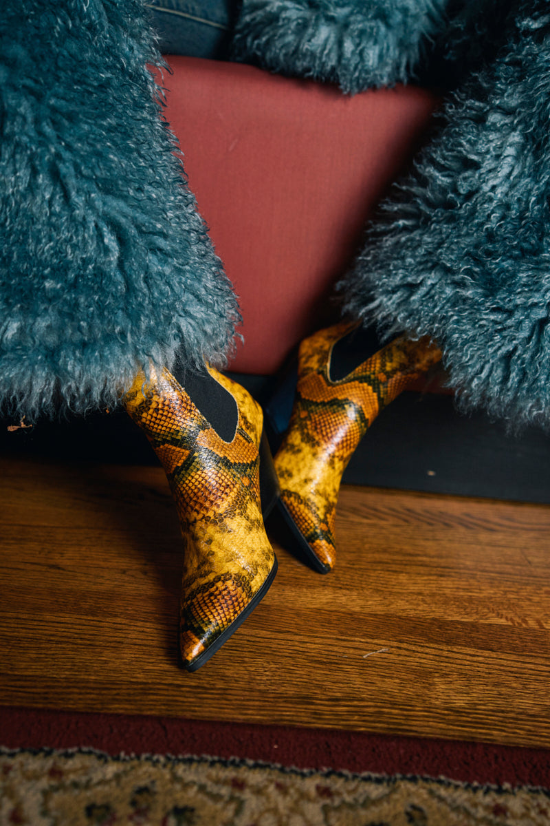 Wearing AZALEA WANG HENDERSON YELLOW CHUNKY BOOTIE and teal shaggy pants, a person sits on a reddish sofa with wooden floor and patterned rug partially visible.