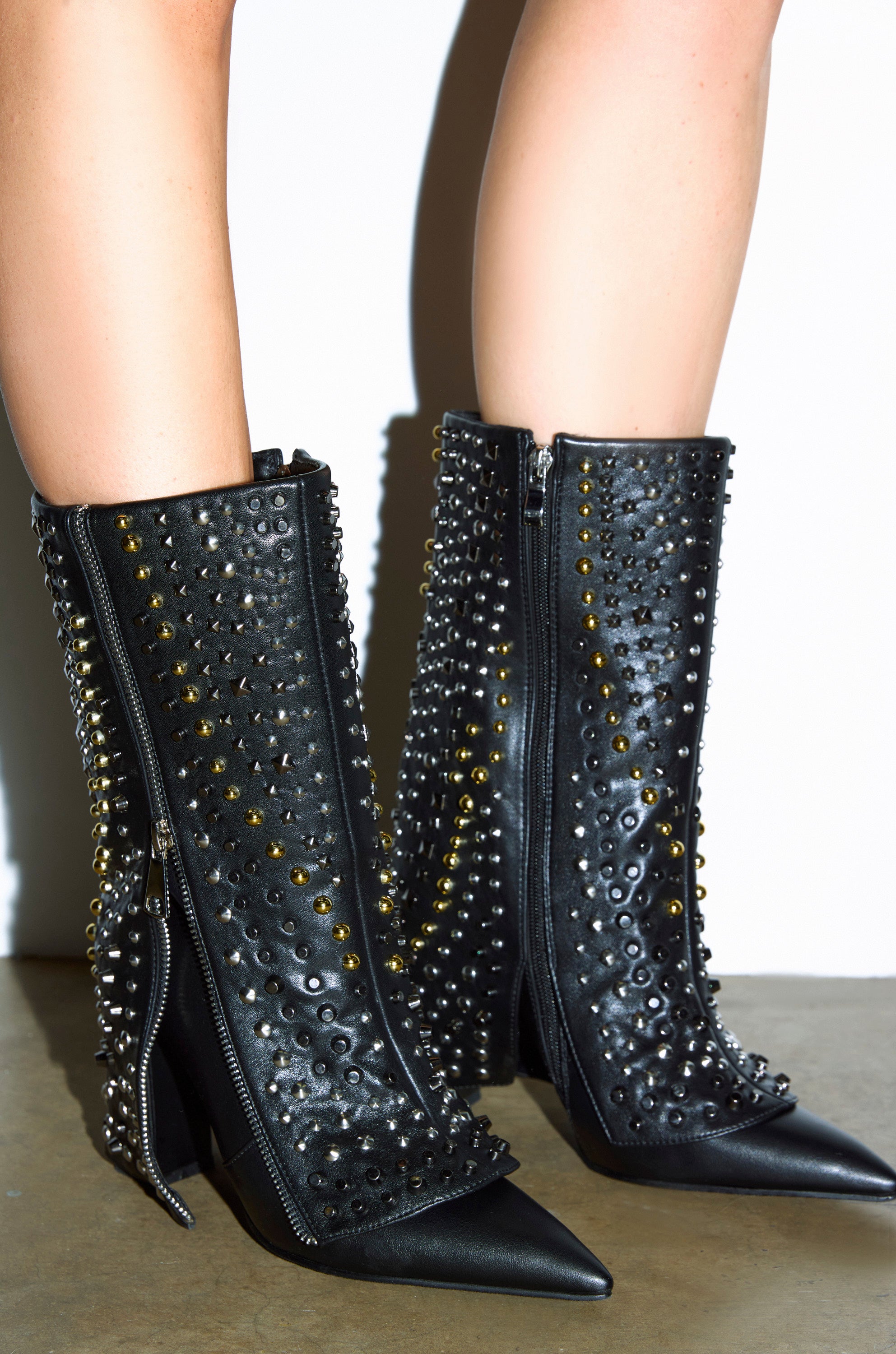 A close-up of a person wearing the AZALEA WANG HENZI BLACK HARDWARE EMBELLISHED CHUNKY FOLD OVER BOOTIE—pointed-toe, knee-high faux leather boots with silver and gold studs, side zippers, and a chunky block heel—styled with bare legs.